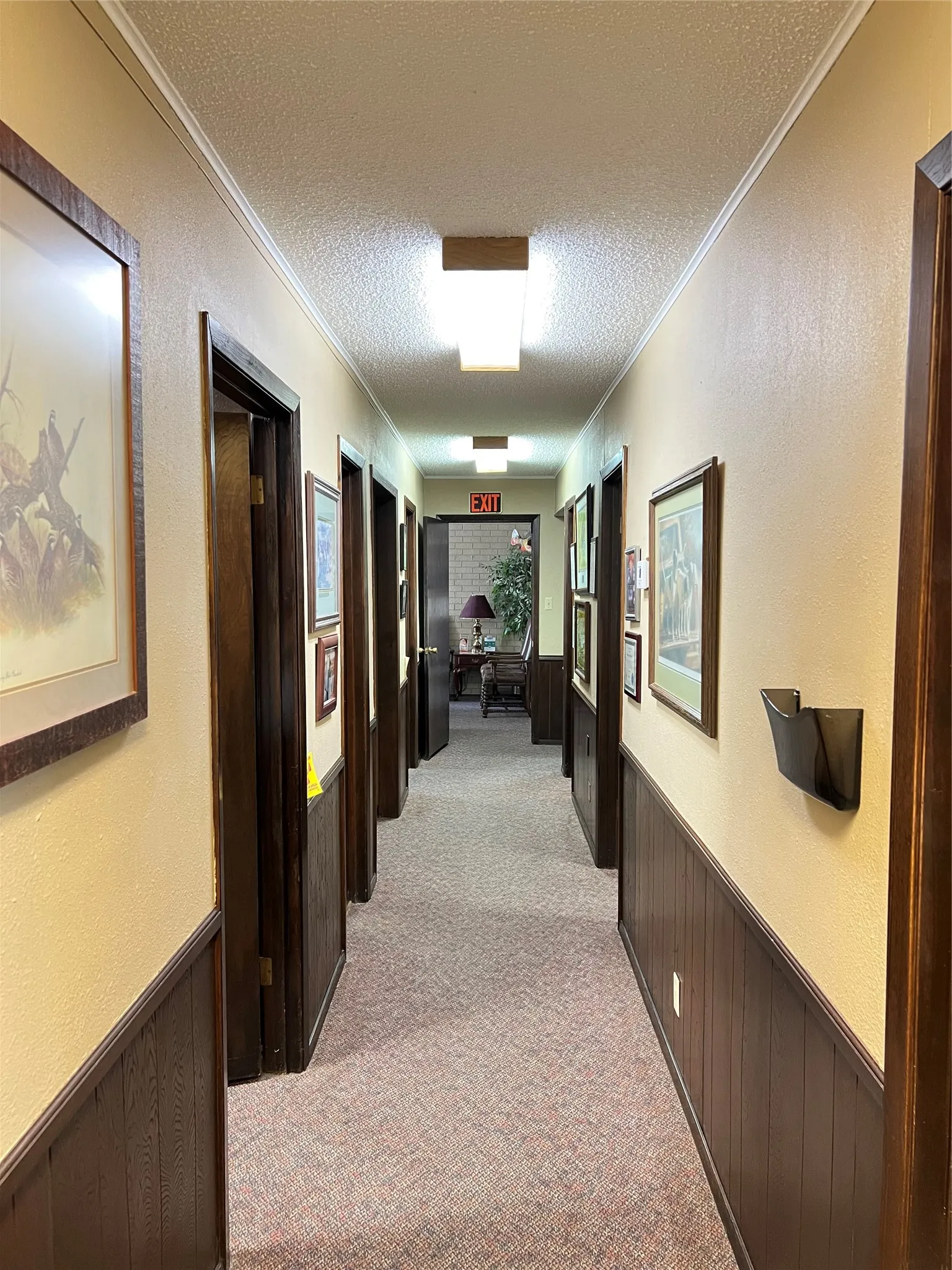 Hallway featuring wainscoting with dark-finish wood trim, multiple door frames with matching wood casing, wall-mounted lighting fixtures, and textured wall surfaces