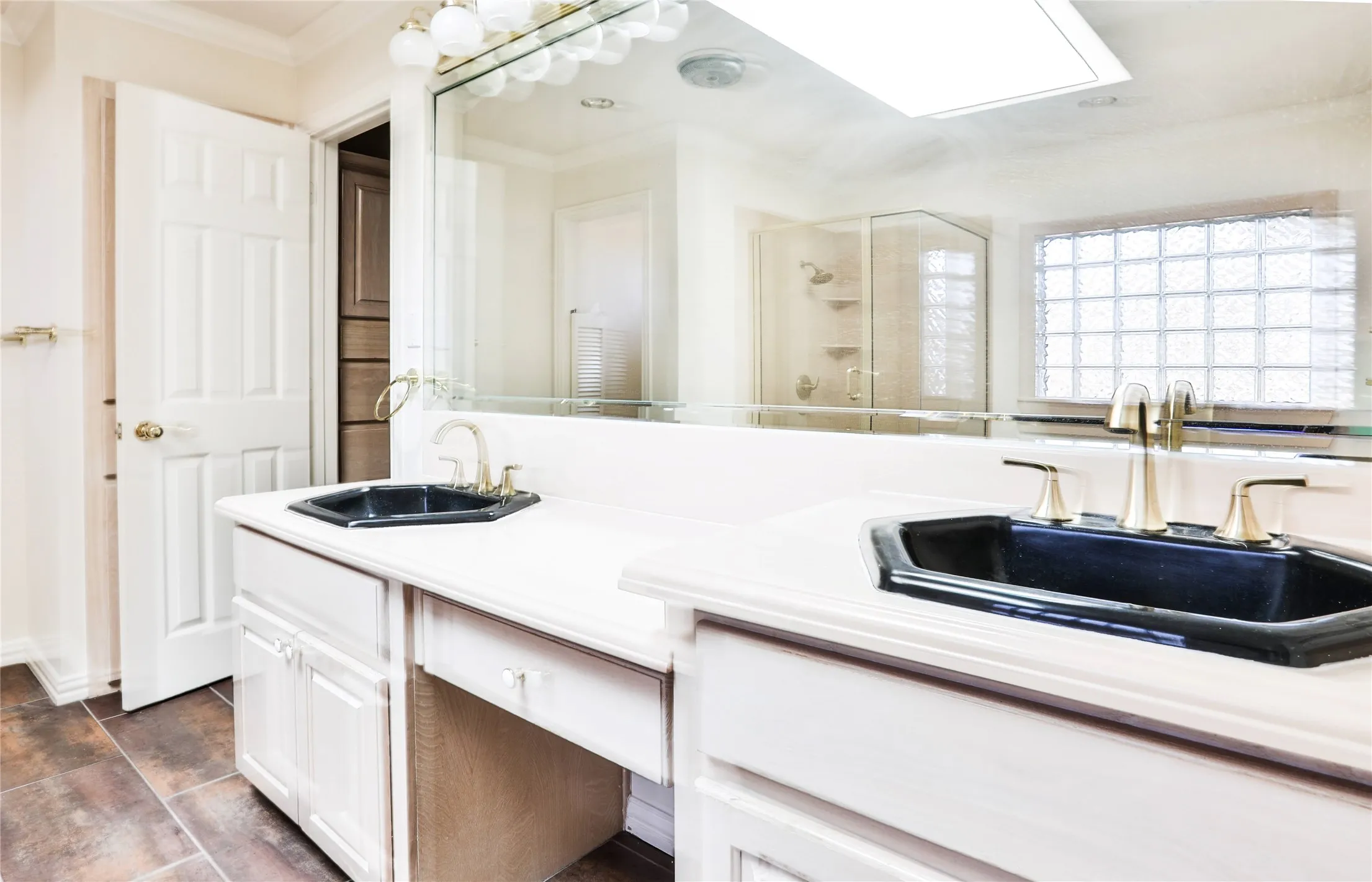 Dual-sink vanity with white cabinetry, light-toned countertop, and recessed black drop-in sinks featuring brass-finish faucets, complemented by a large wall-mounted mirror and glass block window