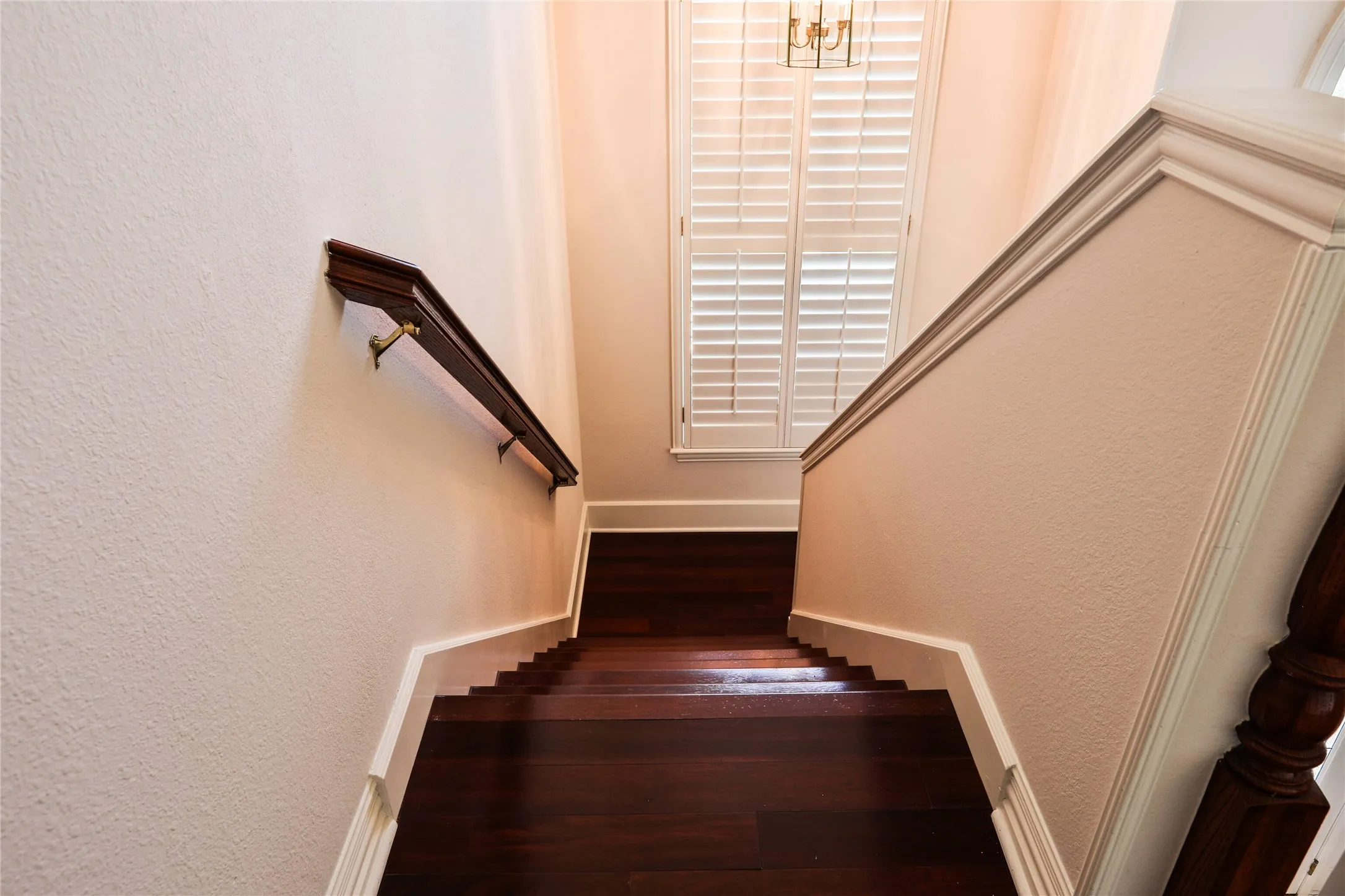 Staircase featuring wood-finish flooring, dark wood handrails with metallic hardware, and plantation shutters