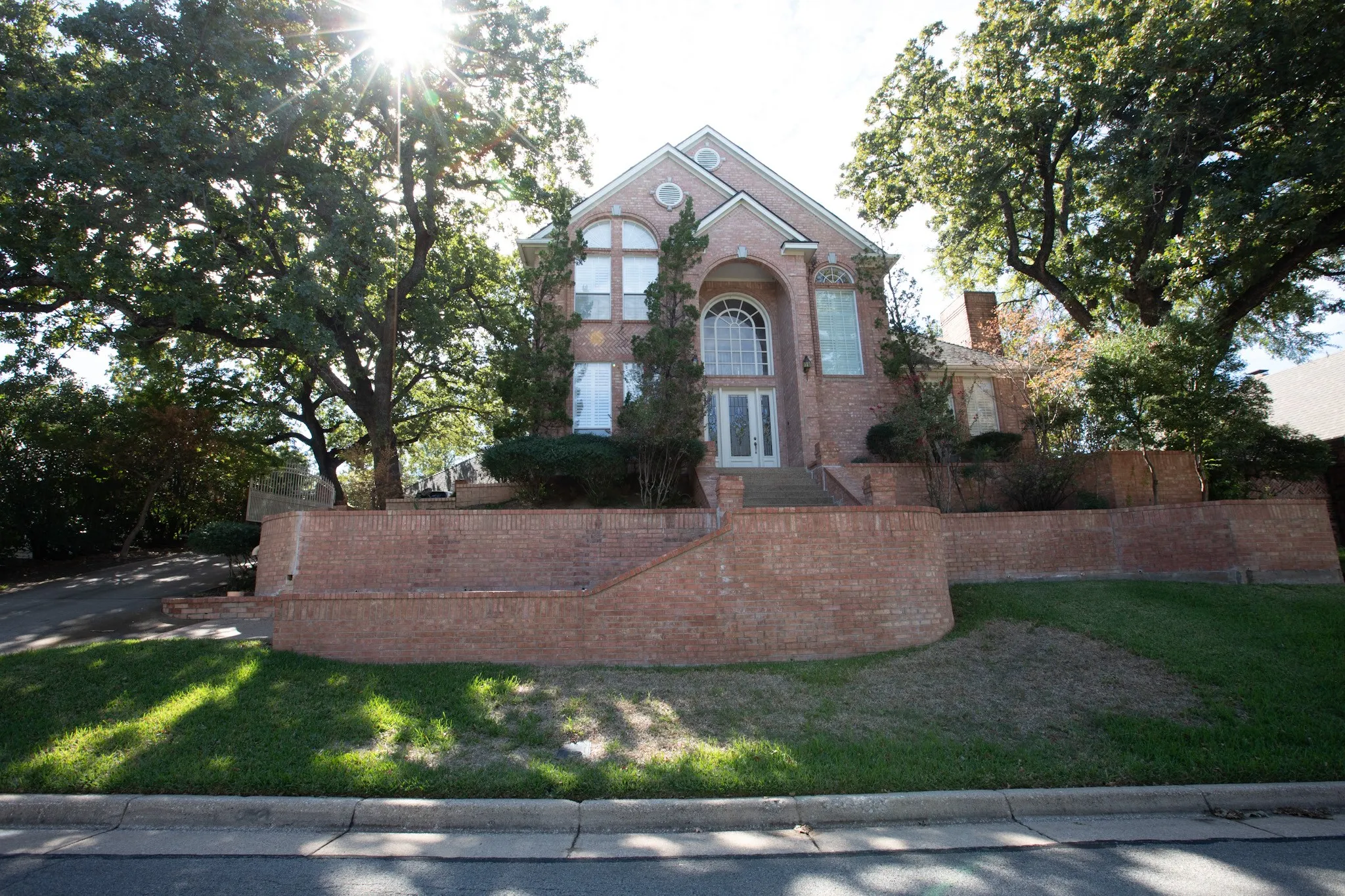 Two-story brick exterior featuring a prominent arched entryway, decorative transom windows, a multi-level terraced brick retaining wall, and mature trees