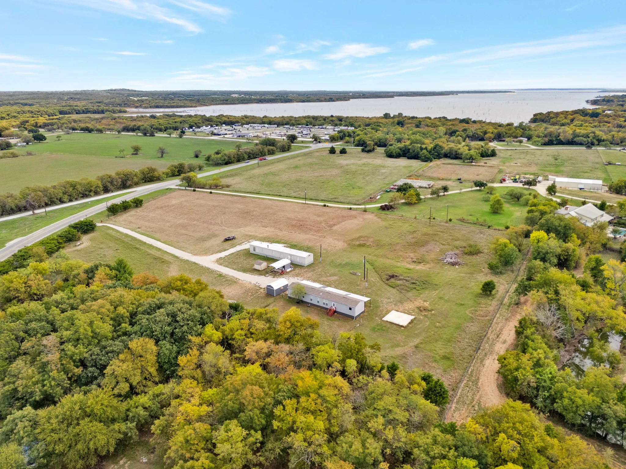 Aerial perspective of a sprawling property featuring expansive open acreage, mature trees, two auxiliary structures, and proximity to a large body of water
