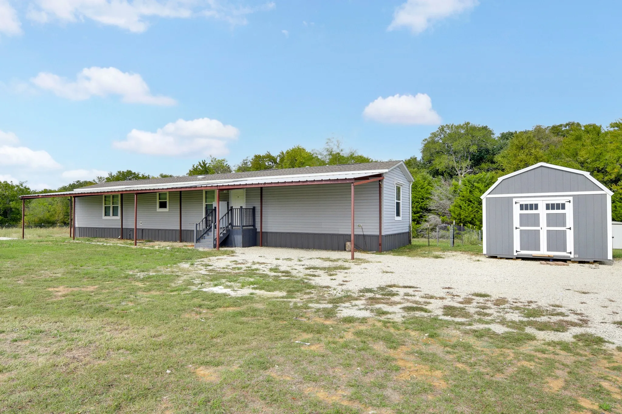 Single-level residence featuring gray horizontal siding, a full-length covered porch, a gravel driveway, and a detached storage structure