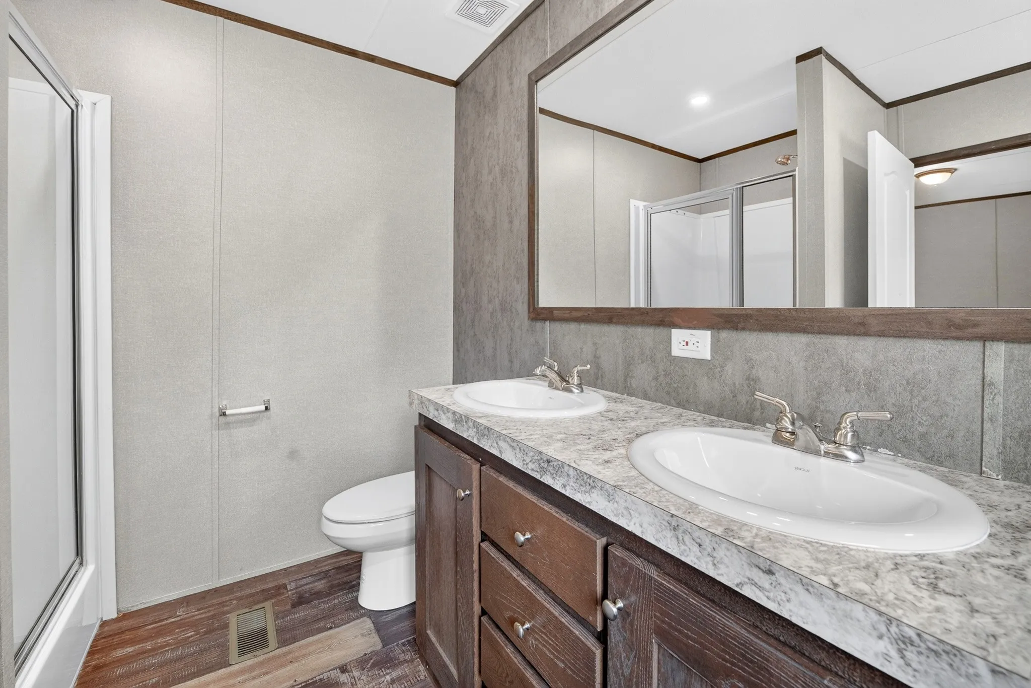 Dual-sink vanity with stone-finish countertop, dark wood-tone cabinetry, large wall-mounted mirror, and glass-enclosed shower stall
