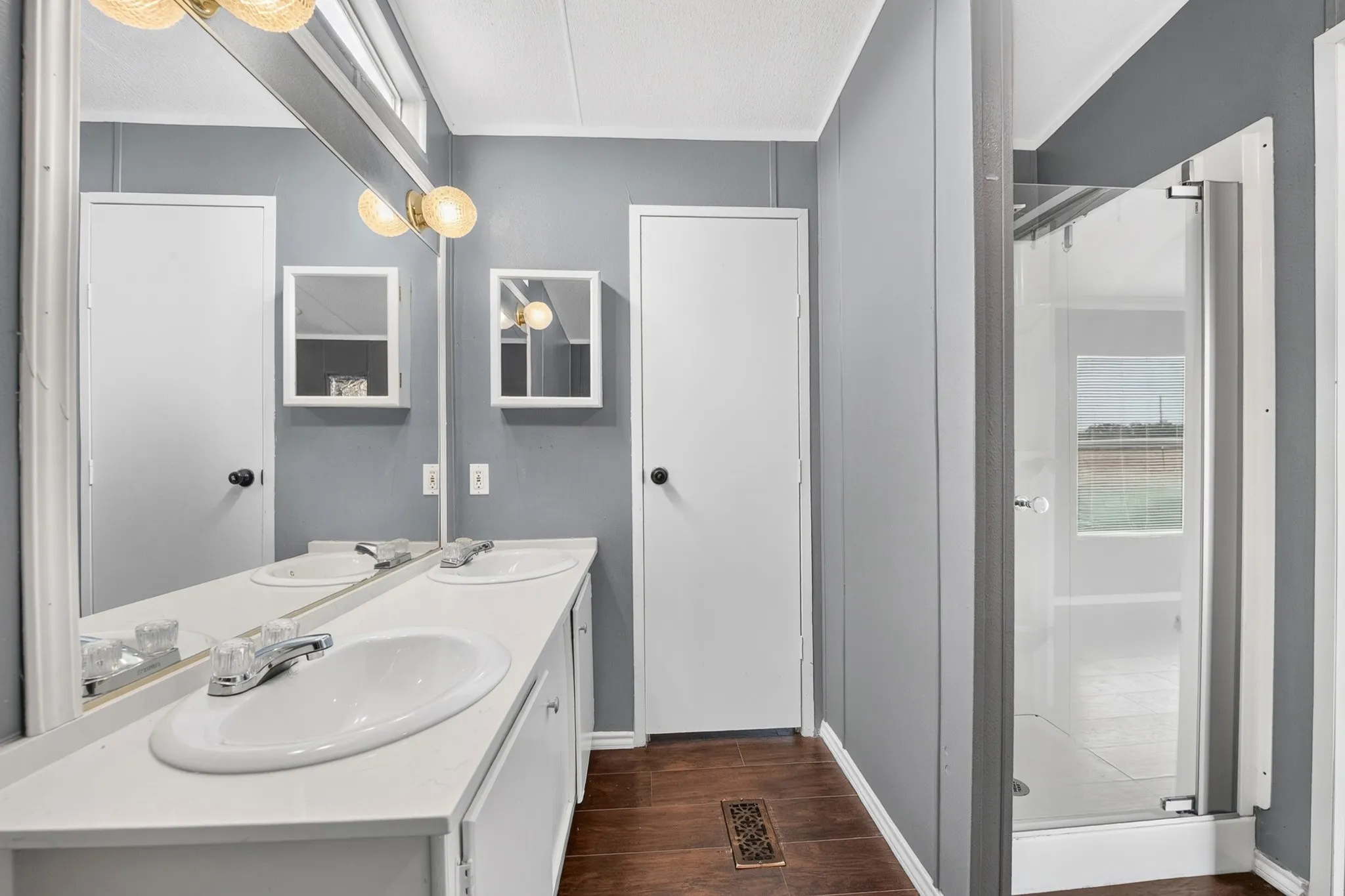 Dual-sink vanity with a white countertop and chrome fixtures, wall-to-wall mirror, two recessed medicine cabinets, and a glass-enclosed shower