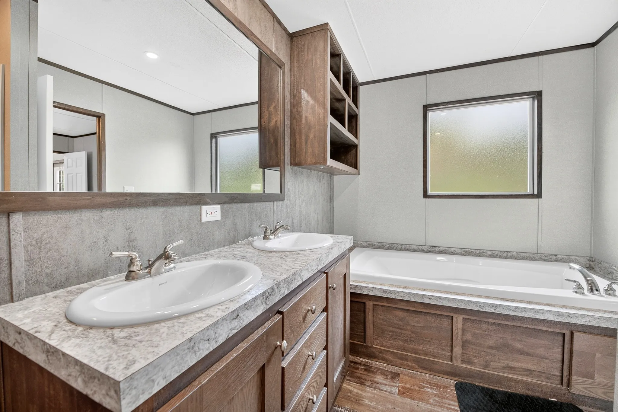 Dual-sink vanity with patterned stone-finish countertop, wood-finish cabinetry, built-in shelving, large wall-mounted mirror, and a soaking tub with matching surround