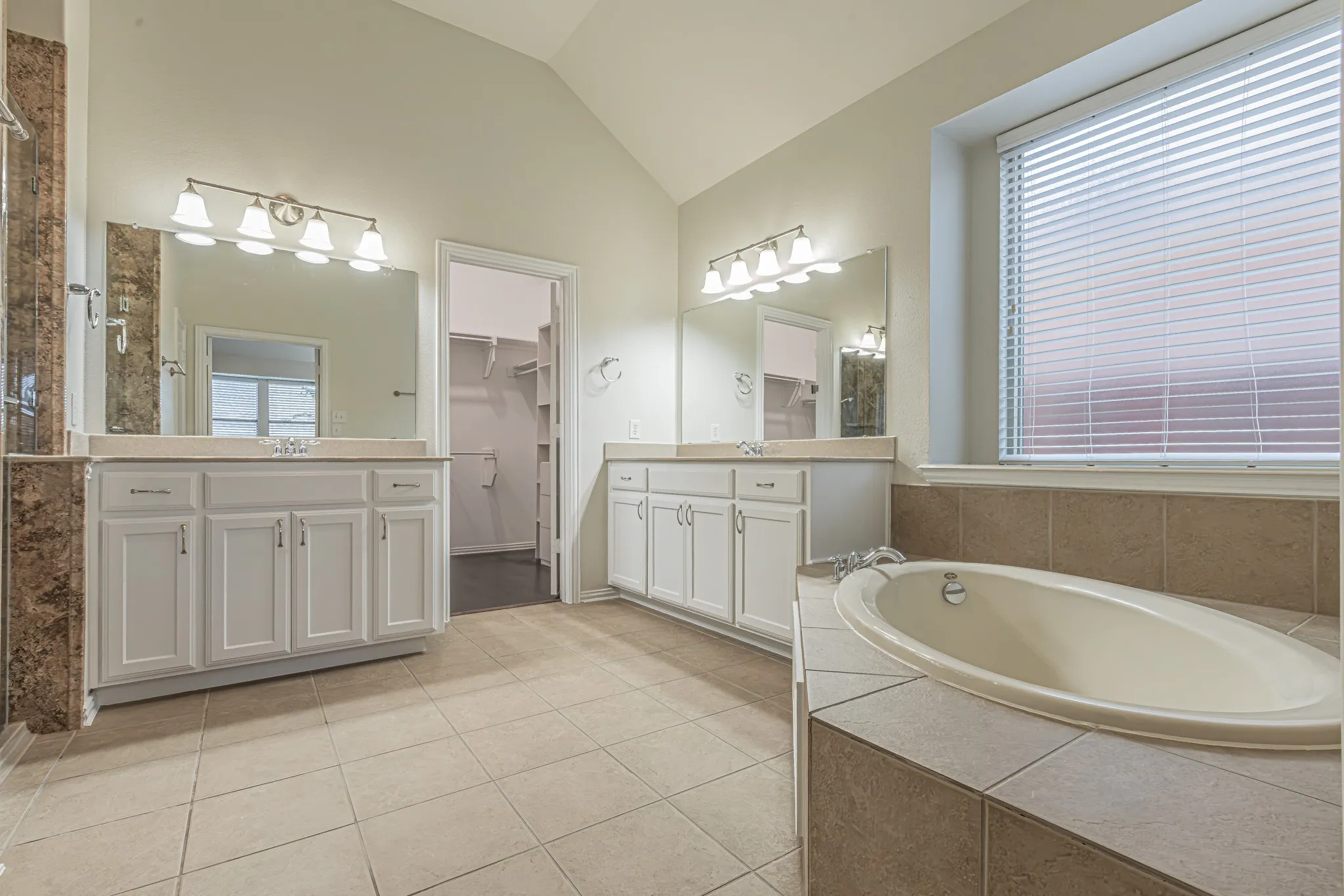 Bathroom featuring a vaulted ceiling, dual white cabinetry vanities with light-toned countertops, a built-in soaking tub with a tiled surround, and large-format tile flooring