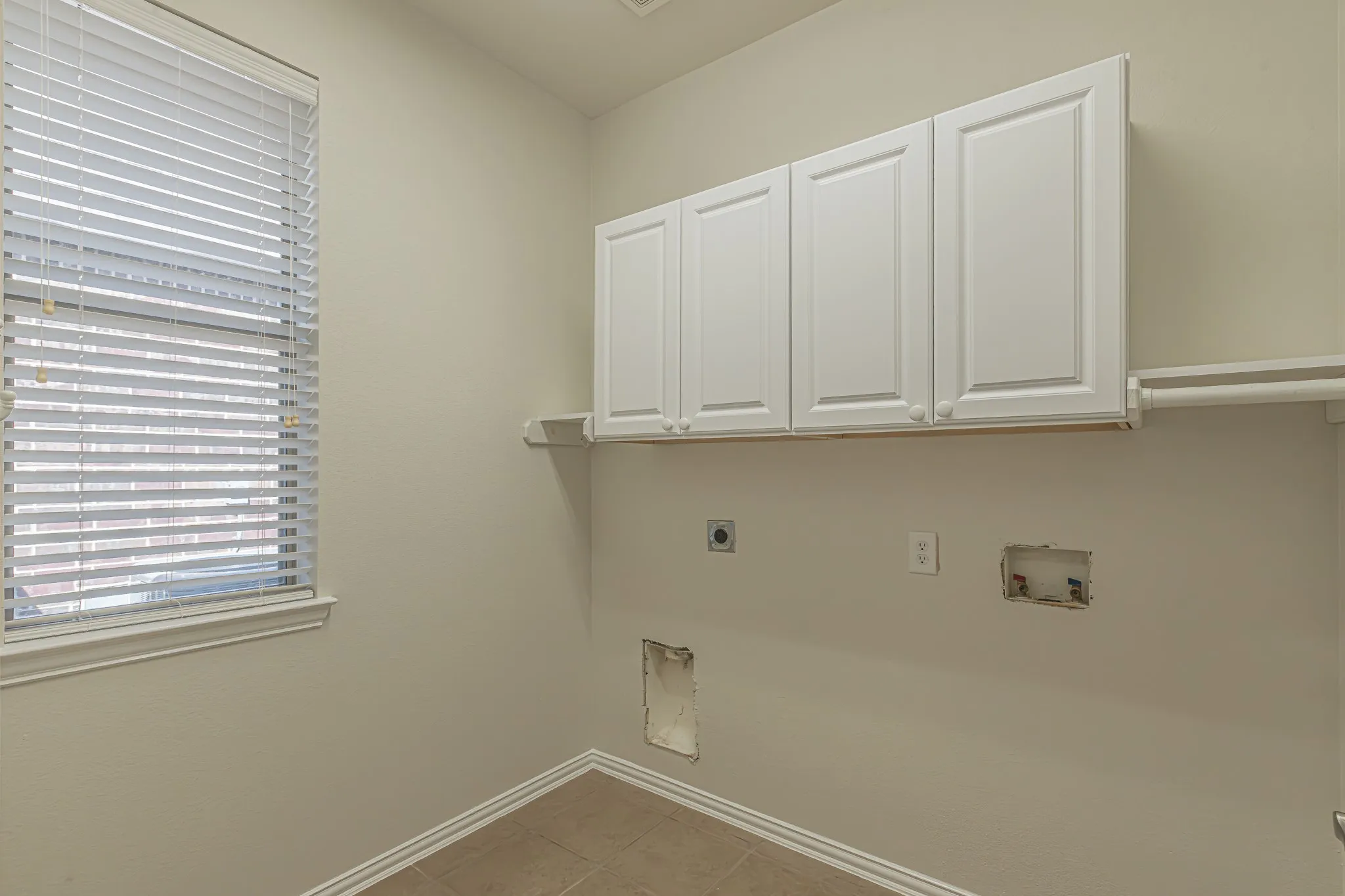 Utility area featuring white shaker-style wall cabinets, a built-in shelf, horizontal slat window blinds, and tile flooring