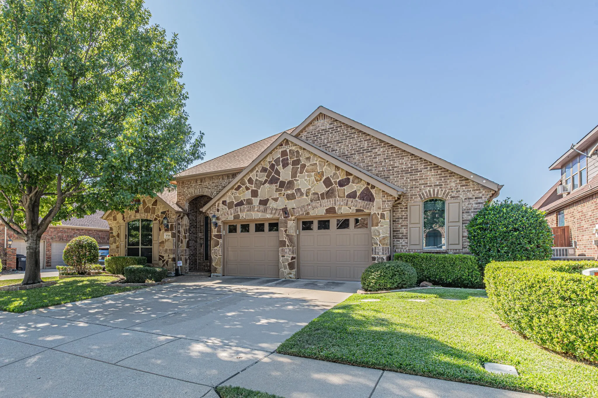 Exterior facade featuring brick and stone masonry, two-car garage, arched window entry, and manicured landscaping