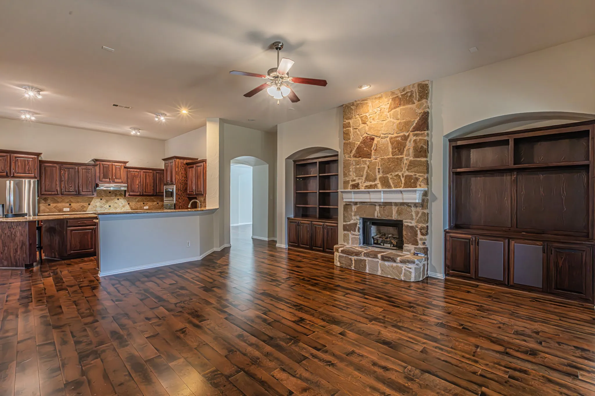 Open-concept area featuring wood-finish flooring, stone-surround fireplace, built-in cabinetry, and kitchen with stained wood cabinetry