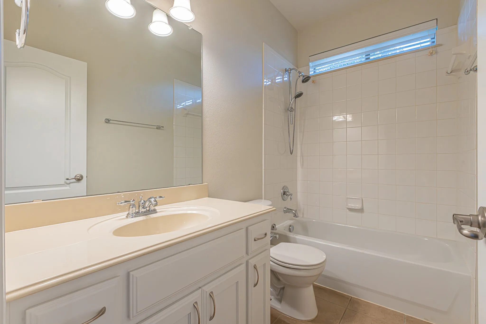 White cabinetry with integrated countertop and vanity sink, full-width wall mirror, three-bulb vanity light fixture, and tile-surround shower and tub combination