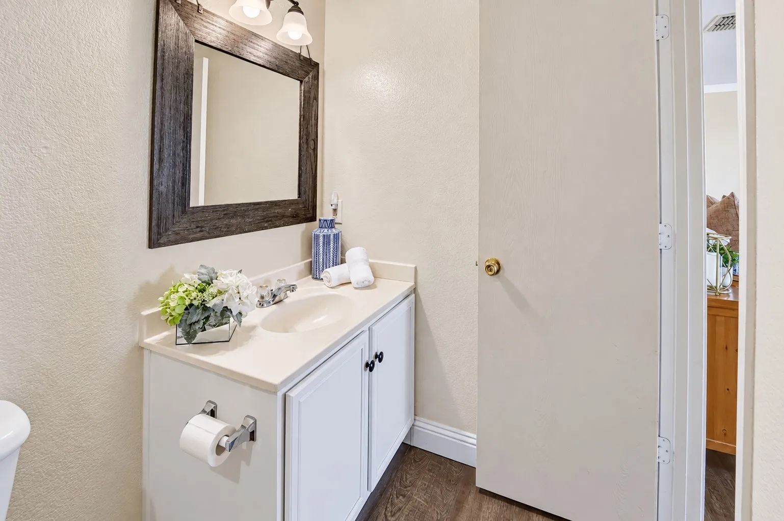 White vanity with a solid-surface countertop and integrated sink, framed rectangular wall mirror, three-light overhead fixture, and wood-finish flooring