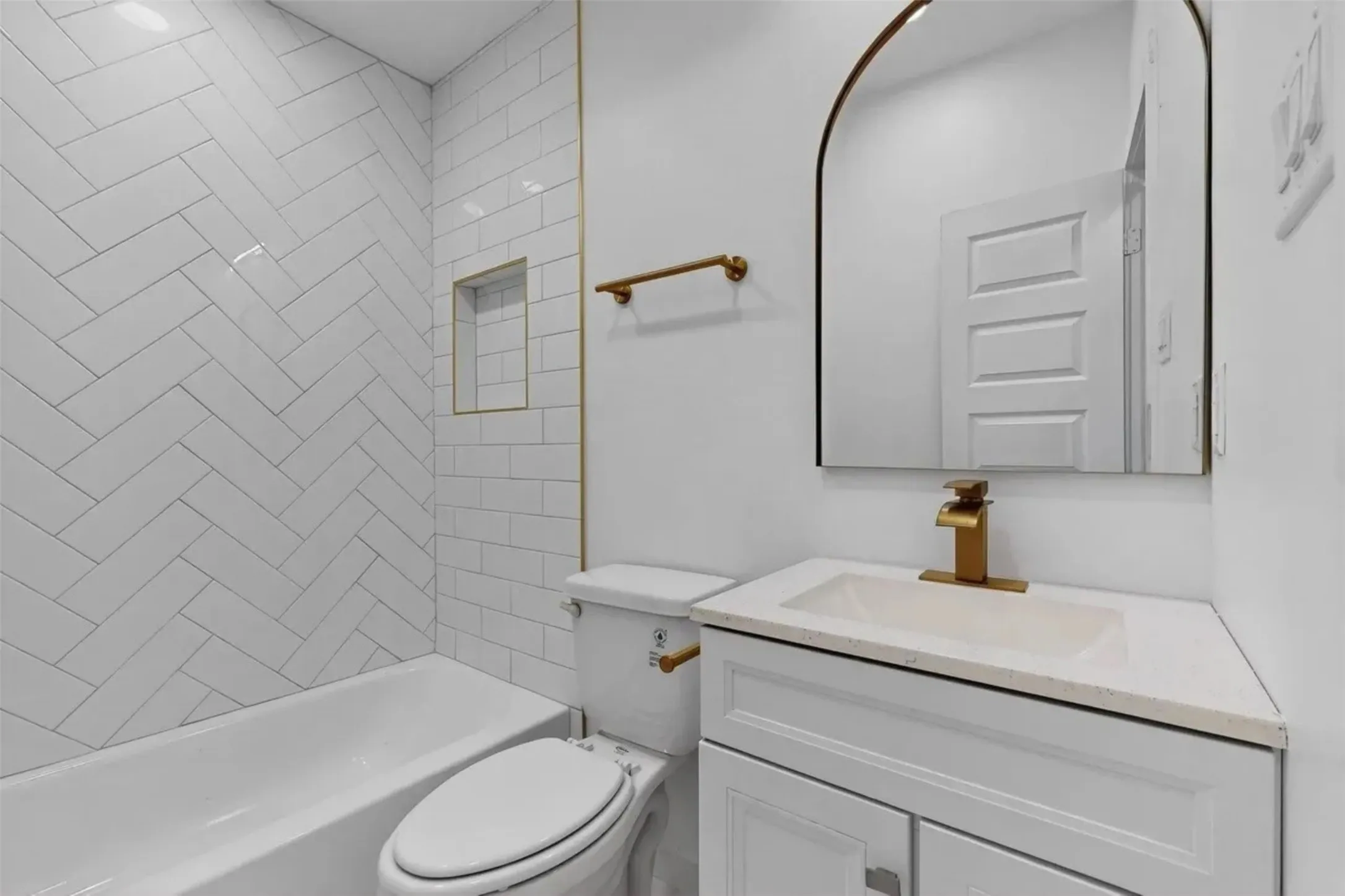 Bathroom featuring a herringbone-patterned tile tub surround, integrated wall niche, arch-top mirror, vanity with stone-composite countertop, and brushed-gold fixtures