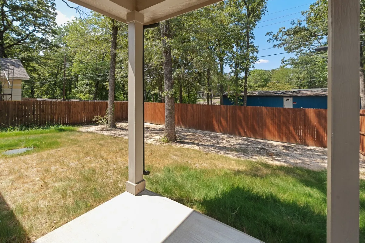 Covered porch featuring concrete flooring and wooden support columns, overlooking a fenced backyard with mature trees