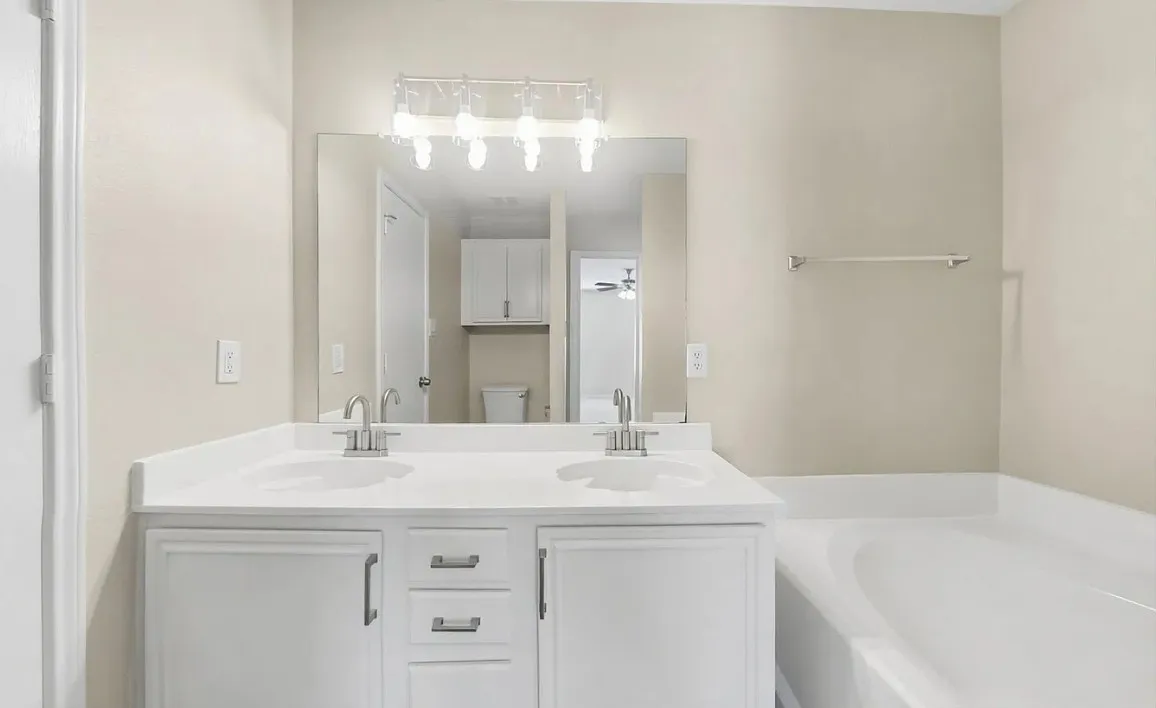 Dual-sink vanity with white cabinetry, solid-surface countertop, chrome fixtures, large rectangular vanity mirror, and built-in soaking tub