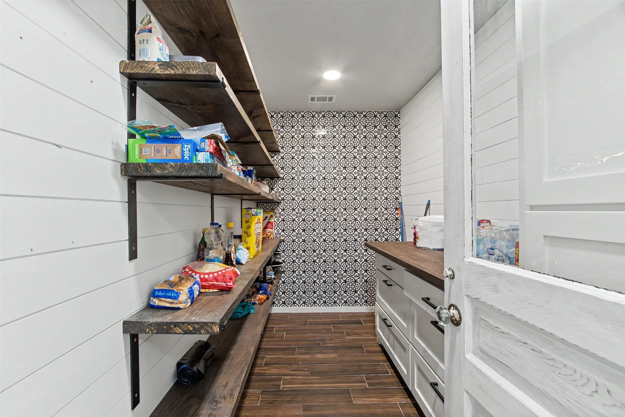 Walk-in pantry featuring open wood-finish shelving, shiplap wall paneling, patterned tile accent wall, wood-finish flooring, and white cabinetry with dark hardware