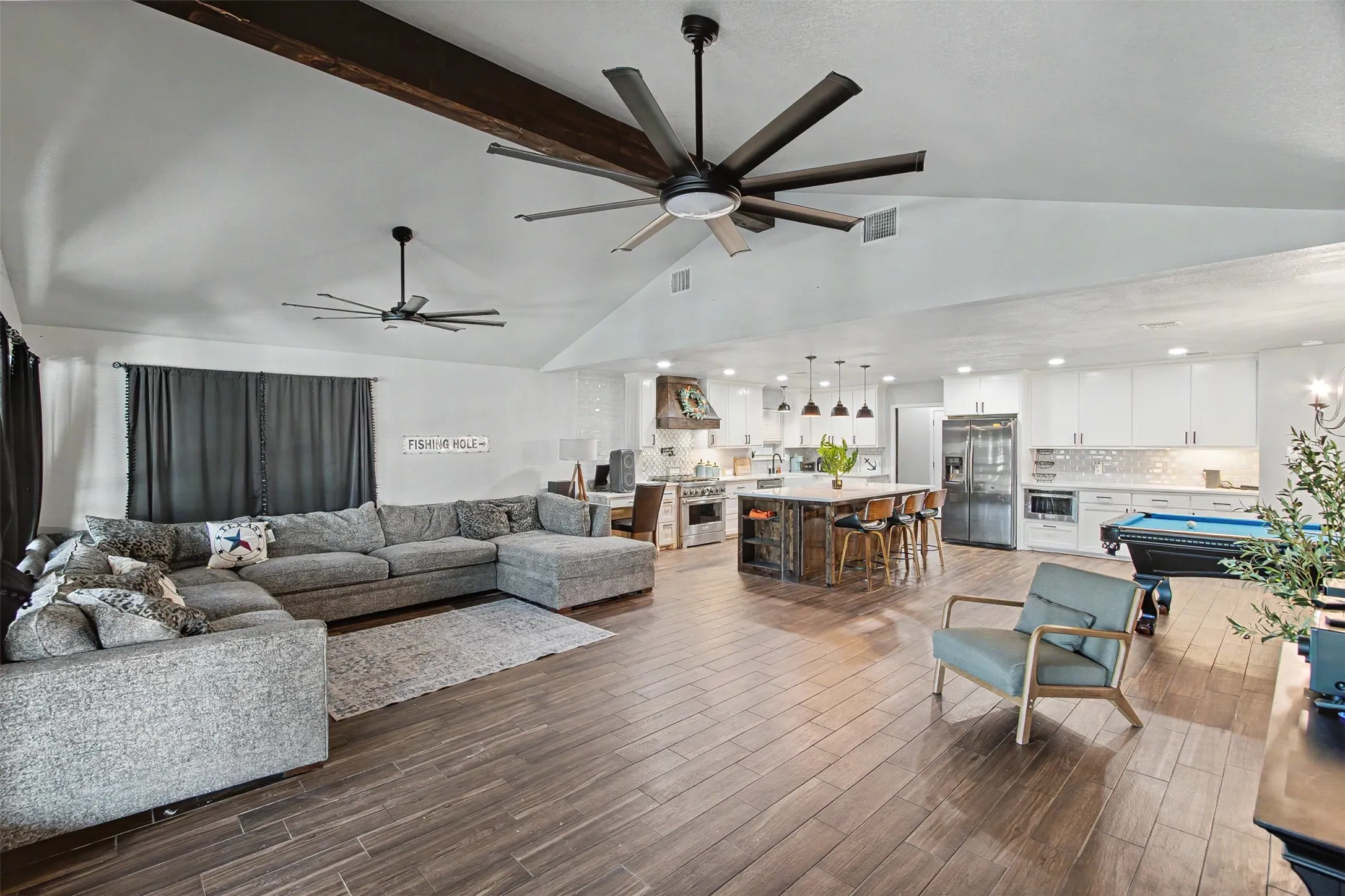 Vaulted ceiling featuring exposed structural beams and dual multi-blade ceiling fans, wood-finish flooring, central kitchen island with bar seating, and white shaker-style cabinetry