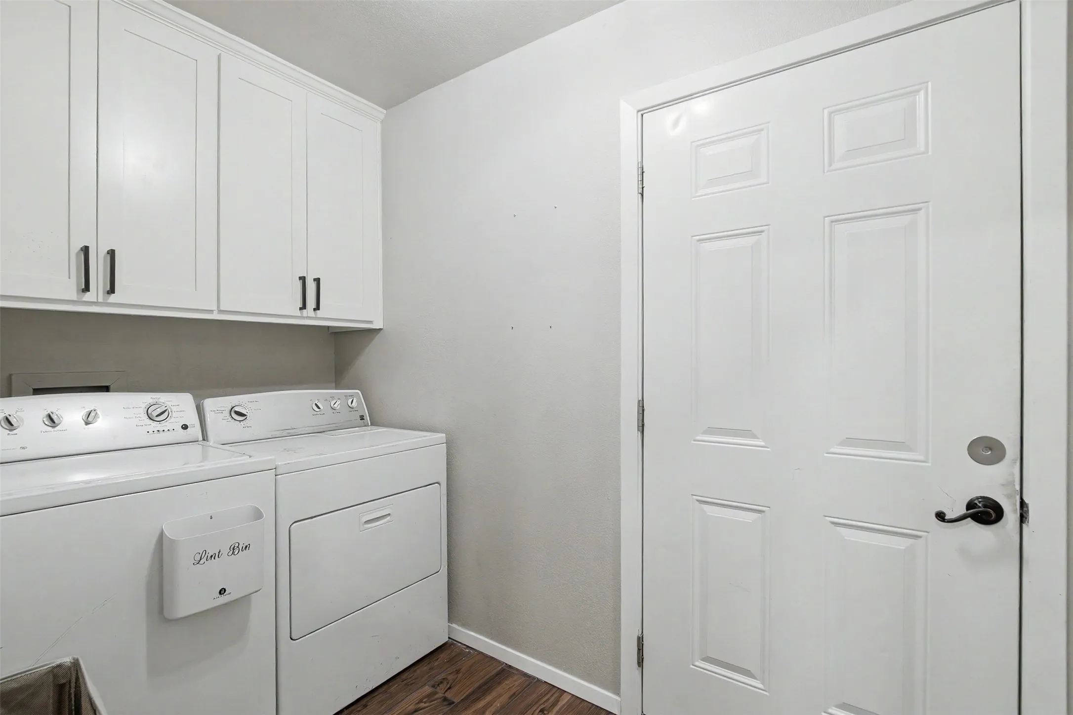 Laundry area featuring white Shaker-style wall cabinetry with matte black hardware, wood-finish flooring, and a six-panel door