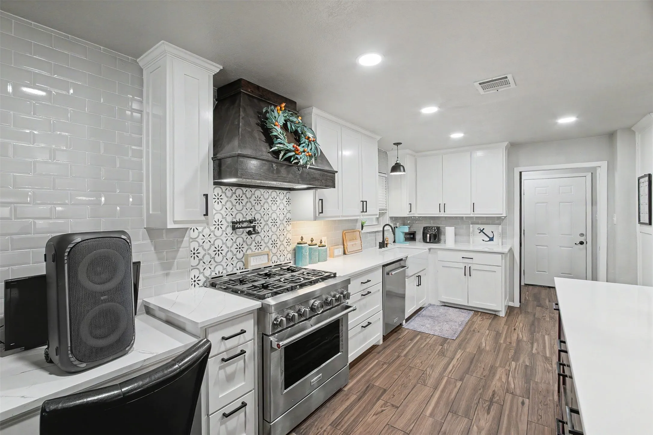 Kitchen featuring white cabinetry, solid-surface countertops, subway tile backsplash with decorative inset, stainless steel gas range with overhead vent hood, and wood-finish flooring