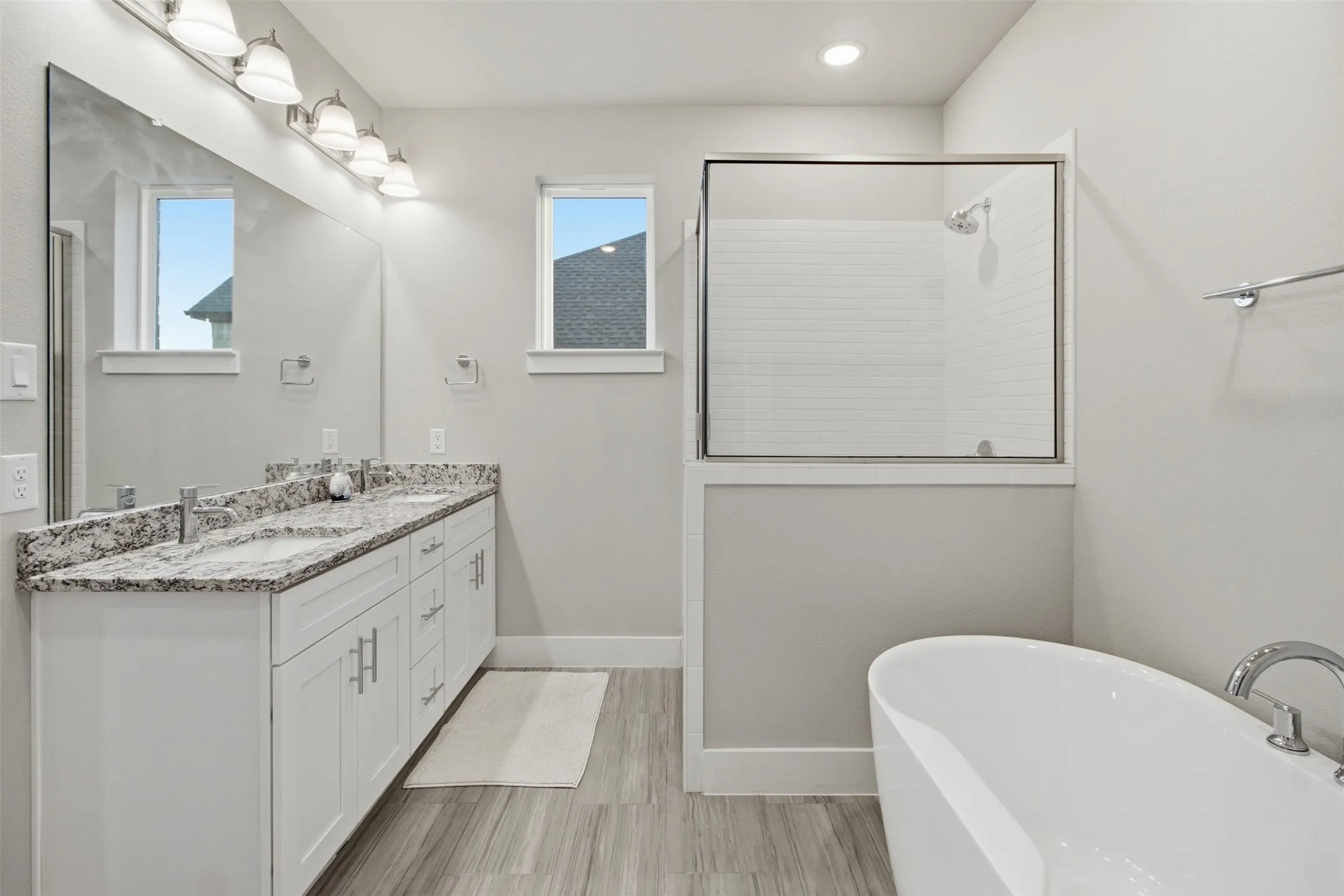 Double vanity featuring stone countertops and integrated sinks, accompanied by a freestanding soaking tub, a tiled glass-enclosed shower, and wood-finish flooring