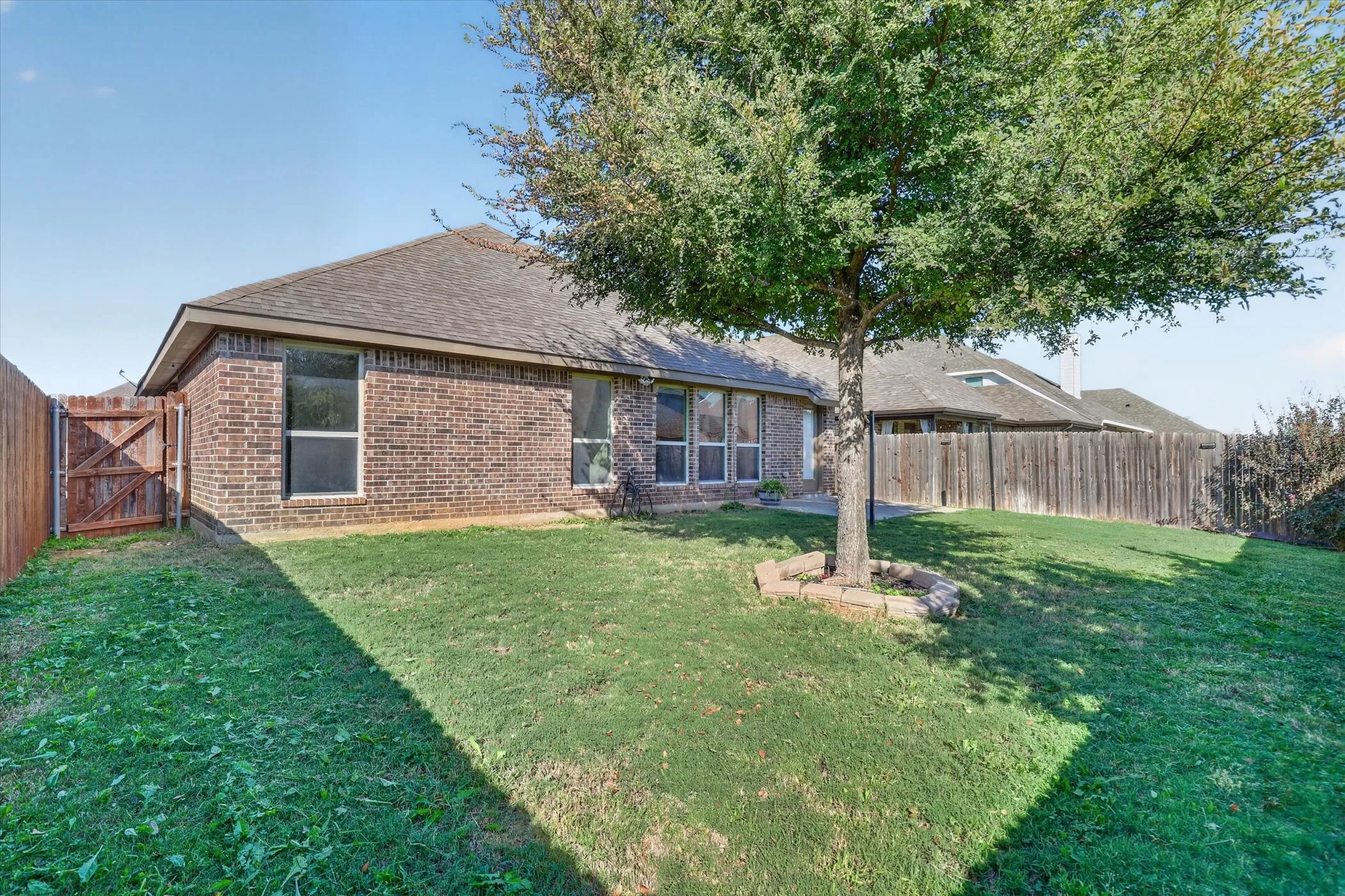 Rear brick exterior featuring a multi-paneled hip roof, wood-fence enclosure, mature landscaping, and stone tree surround