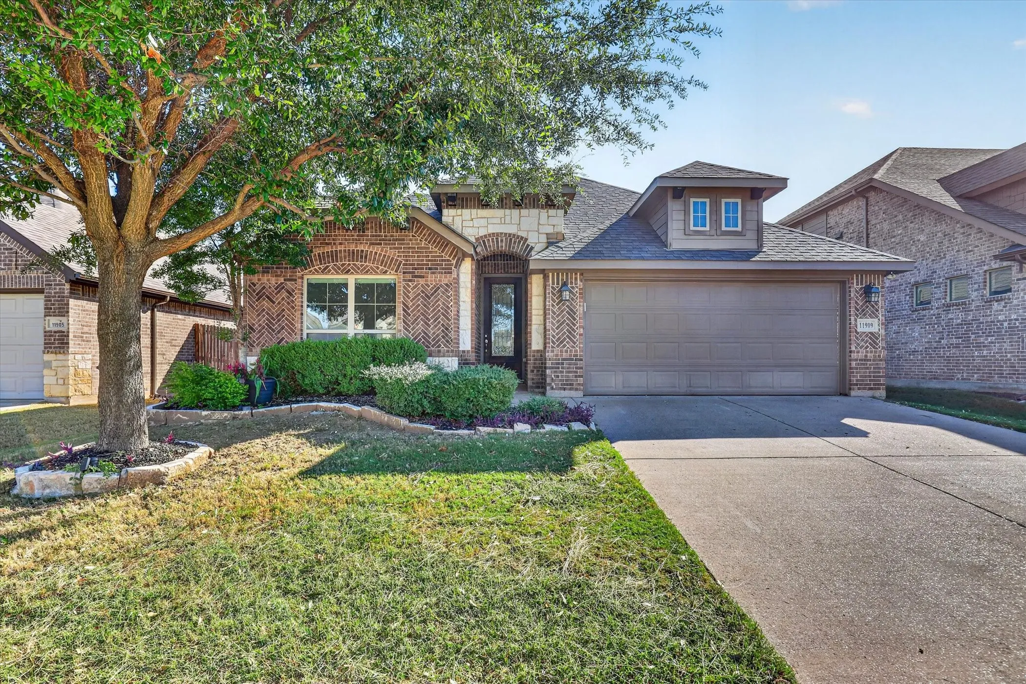 Brick and stone facade, arched entryway, shingle roof, concrete driveway, and mature landscaping