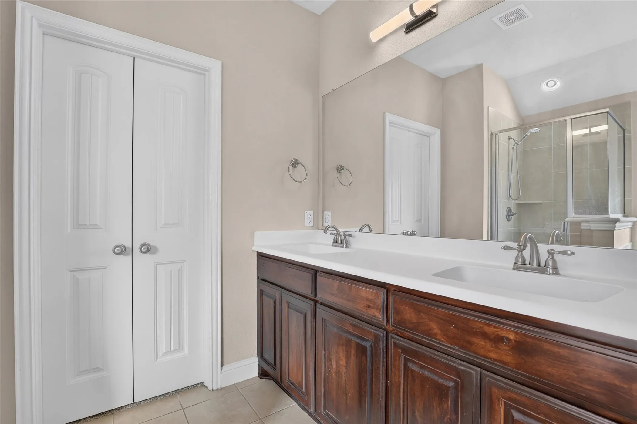 Double-vanity cabinet featuring wood-finish cabinetry, solid-surface countertop with integrated dual sinks, full-width mirror, and walk-in shower enclosure