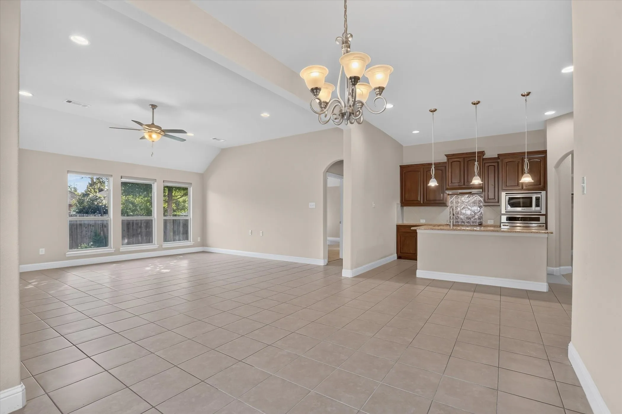 Vaulted ceiling with a central ceiling fan, neutral tile flooring, arched doorways, a traditional chandelier, and kitchen cabinetry featuring built-in stainless steel appliances