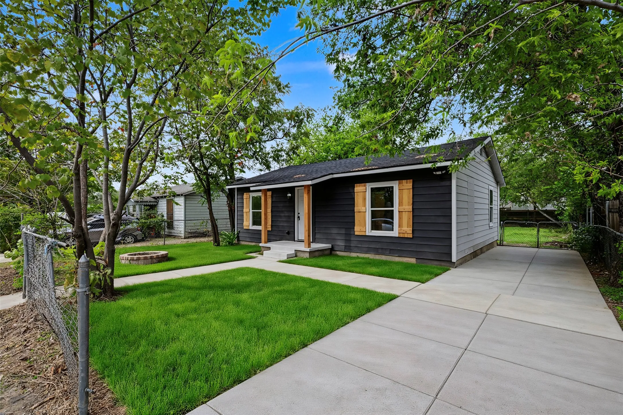 Single-story exterior featuring dark lap siding, wood-tone shutters, a concrete driveway, and a front lawn with a circular stone fire pit