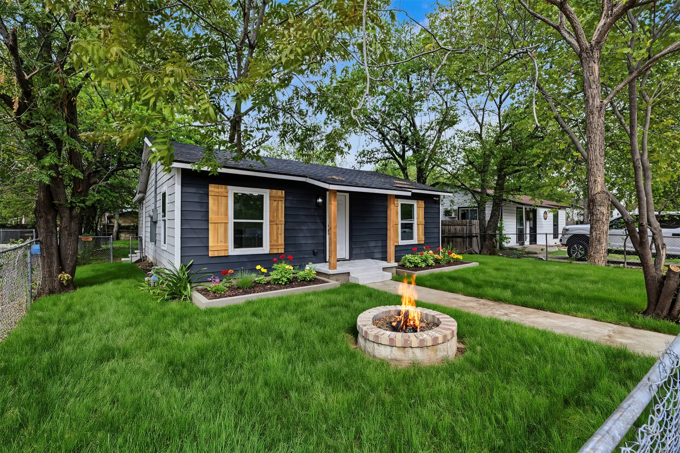 Single-story residence featuring horizontal lap siding, decorative wood shutters, a concrete front stoop, and a stone-encased circular fire pit set within a landscaped lawn