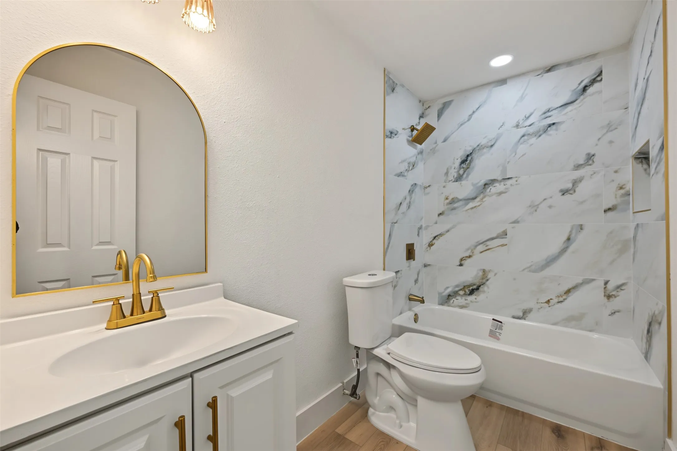 Bathroom featuring a white vanity cabinet with gold-tone hardware, a gold-framed arched mirror, gold-tone faucet, stone-patterned tile shower surround, and wood-finish flooring