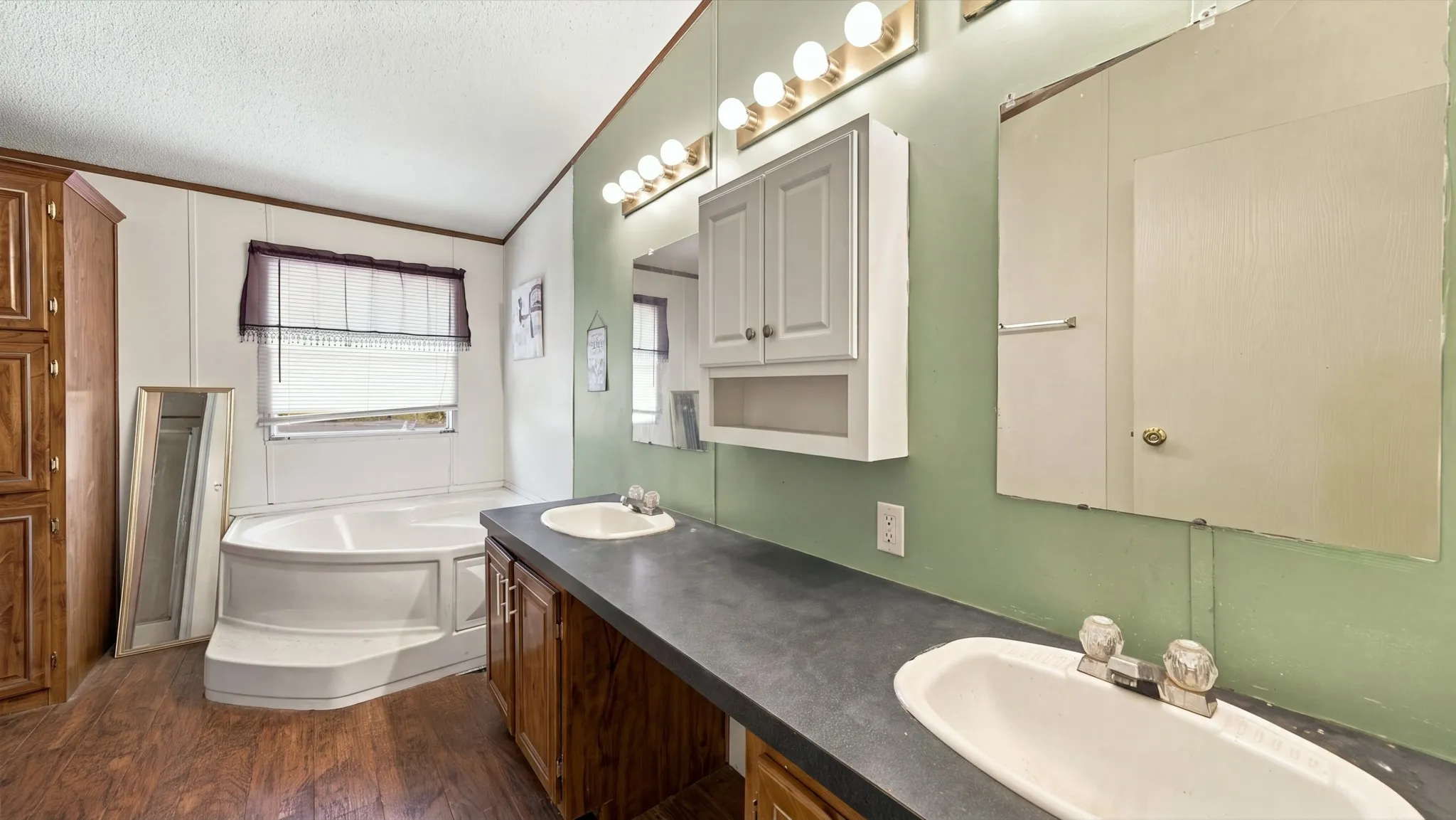 Dual-sink vanity with dark-toned countertops and wood-finish cabinetry, corner soaking tub, wall-mounted medicine cabinet, and dual vanity lighting fixtures