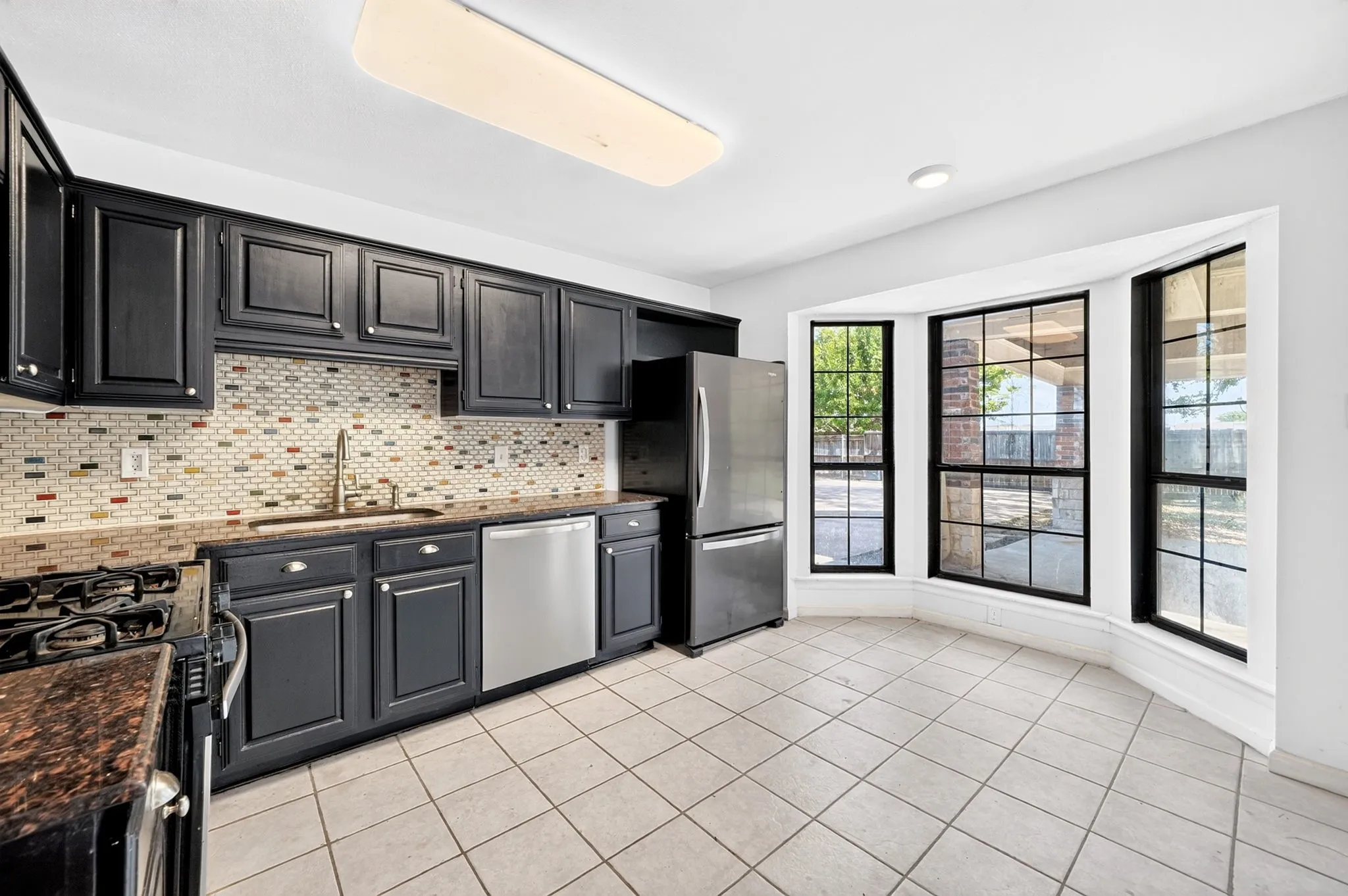 Kitchen featuring dark-finish shaker cabinetry, stone countertops, a multi-tone mosaic tile backsplash, stainless steel appliances, and a bay window