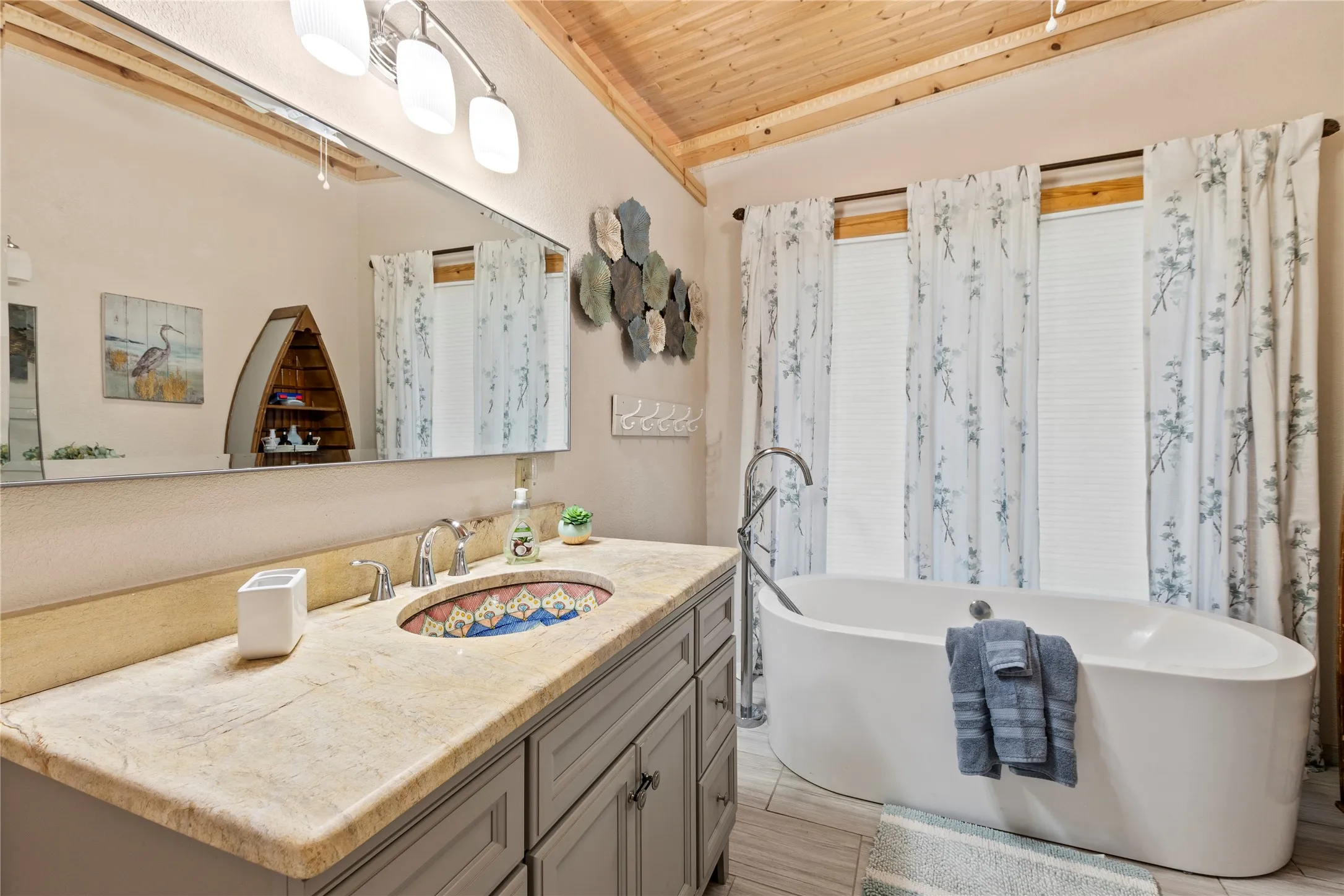 Freestanding soaking tub with a floor-mounted faucet, stone countertop with an integrated patterned basin, grey cabinetry, and wood-paneled ceiling