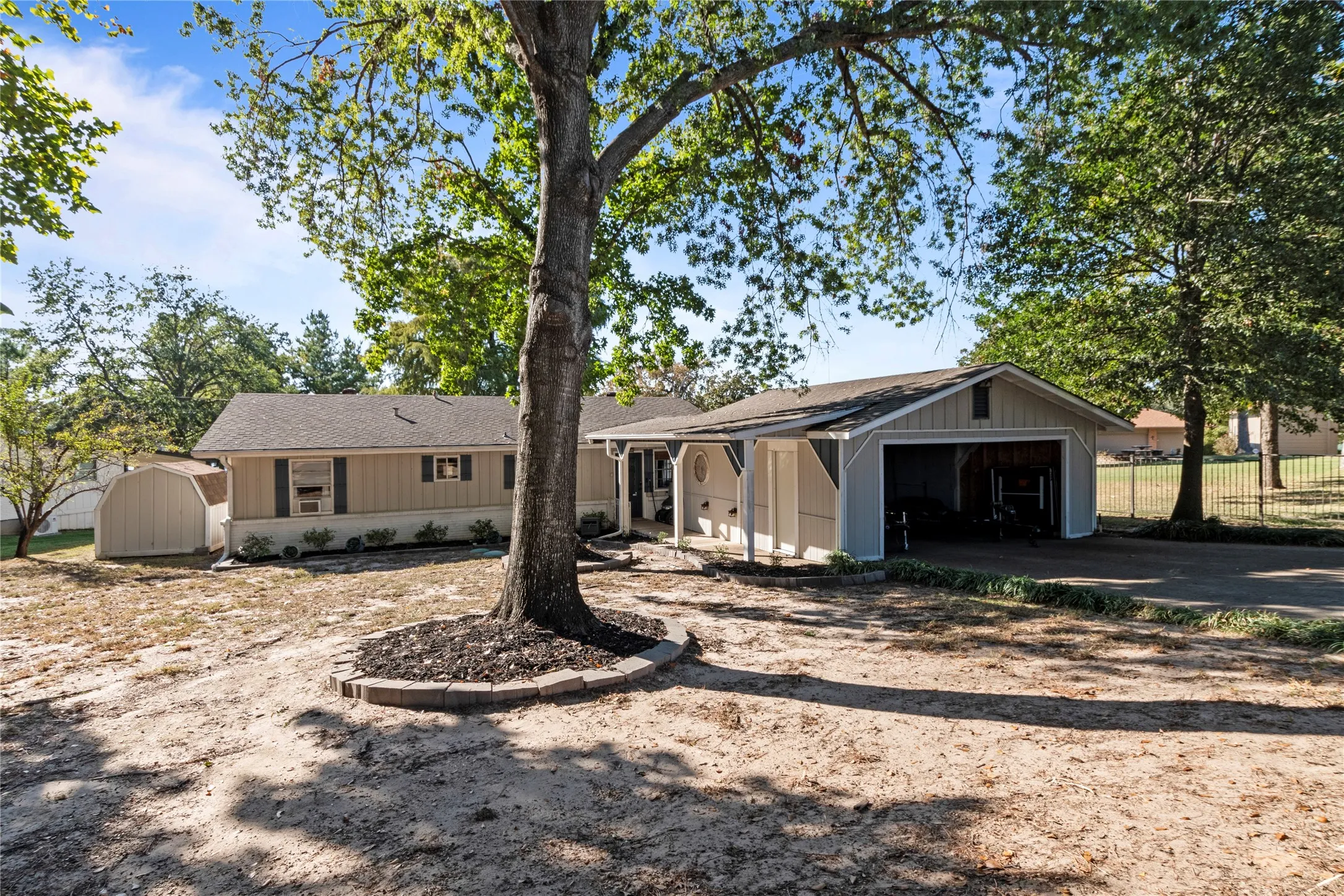 Single-story exterior featuring vertical siding, attached carport, detached storage shed, and stone tree surround