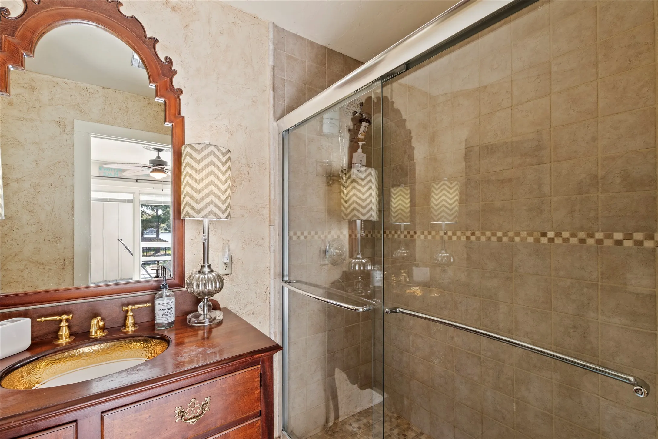 Bathroom featuring a wood vanity with a recessed basin, gold-tone faucet hardware, arched wall mirror, and a walk-in shower enclosure with tile surround