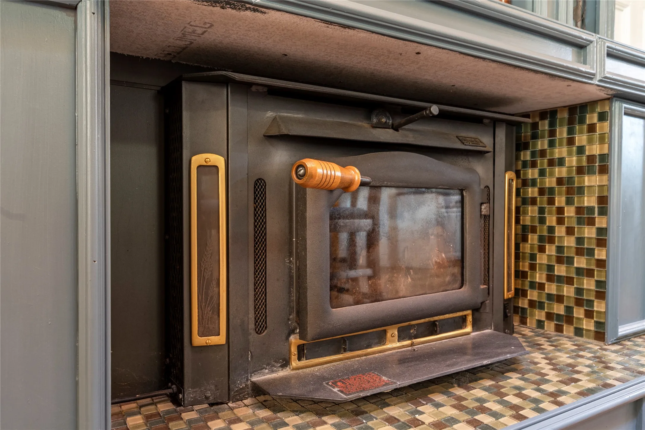 Wood-burning stove insert featuring a matte black finish, brass accents, a glass viewing panel, and a wood-toned handle, framed by mosaic tile surfacing and custom cabinetry