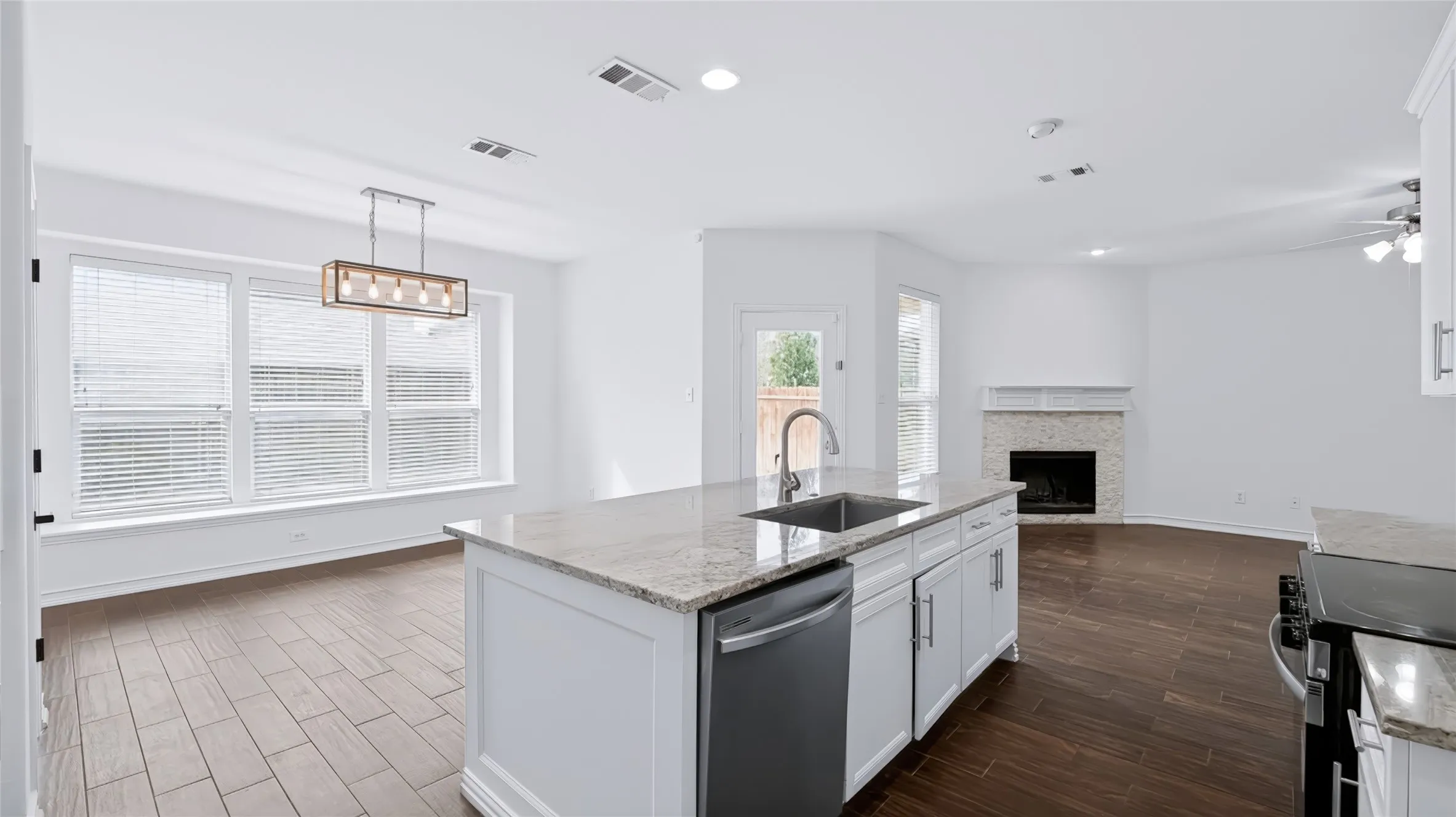 Kitchen island with stone countertop, undermount stainless steel sink, wood-finish tile flooring, linear pendant lighting, and fireplace with stone-tiled surround