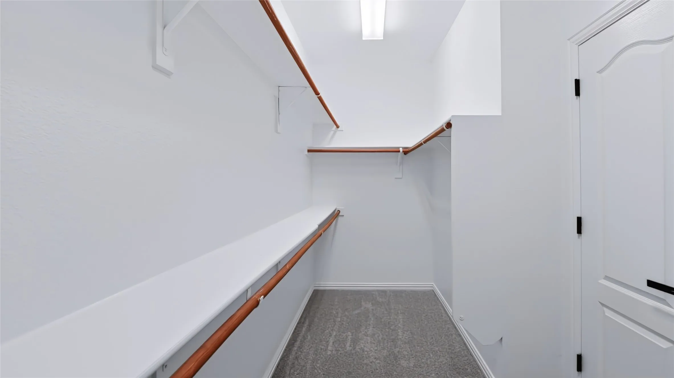 Storage space featuring built-in wood-finish shelving, wall-mounted hanging rods, grey carpeting, and a flush-mount overhead light fixture