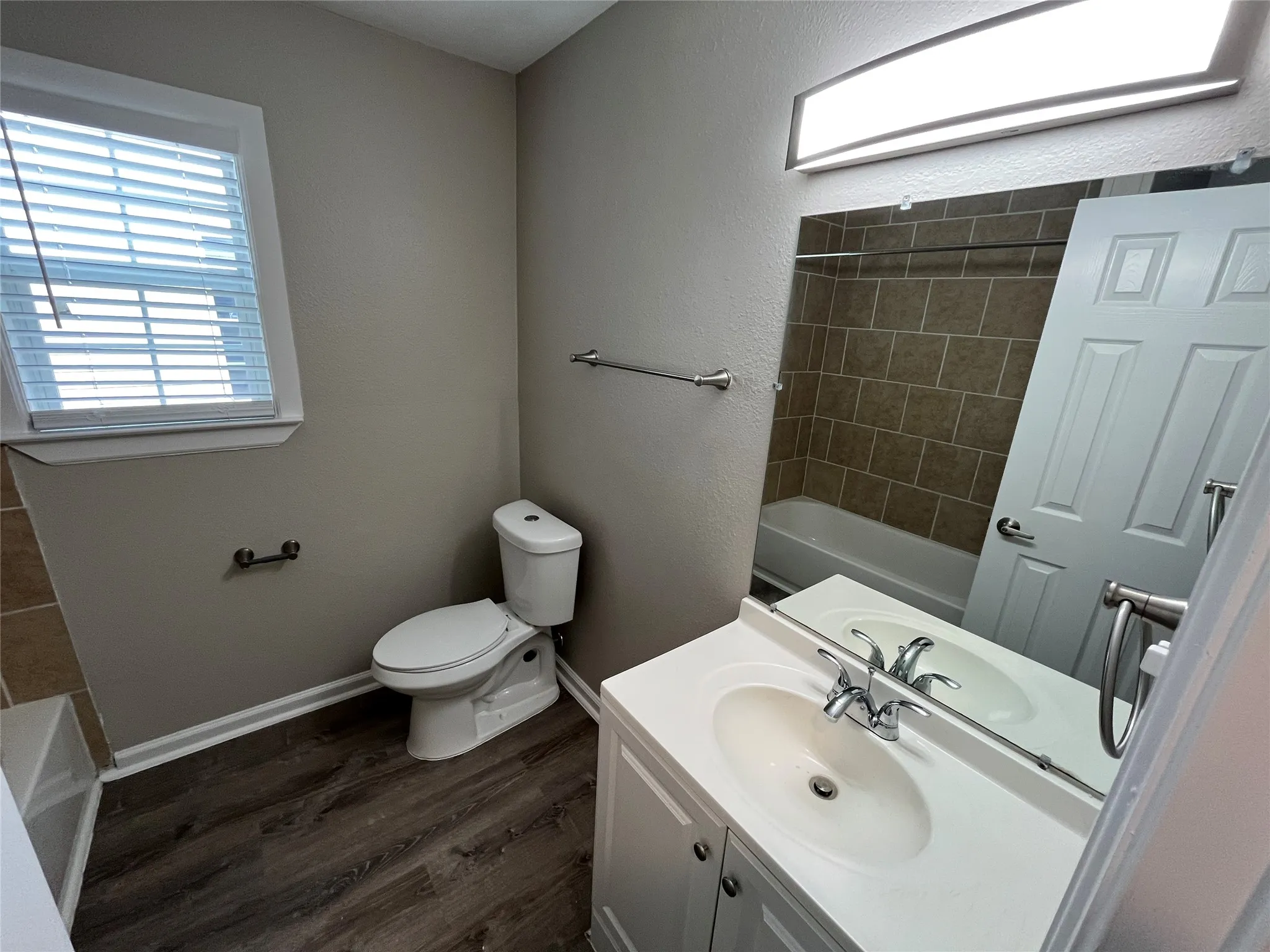 Bathroom featuring a white vanity cabinet, wood-finish flooring, a wall-mounted vanity mirror, a rectangular tub enclosure with tile surround, and brushed nickel hardware