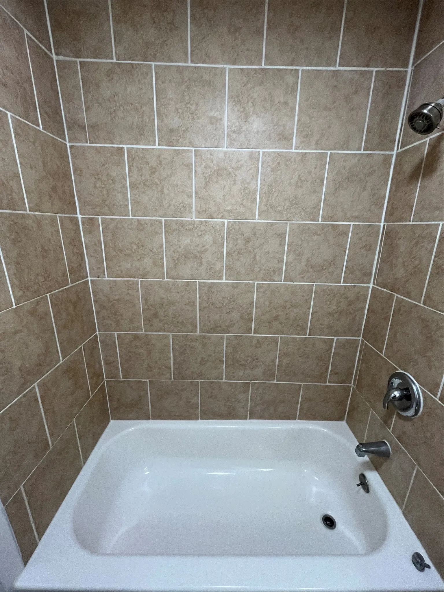 Bathtub surround featuring square, neutral-toned tile with contrasting grout lines, chrome showerhead, and matching chrome faucet assembly