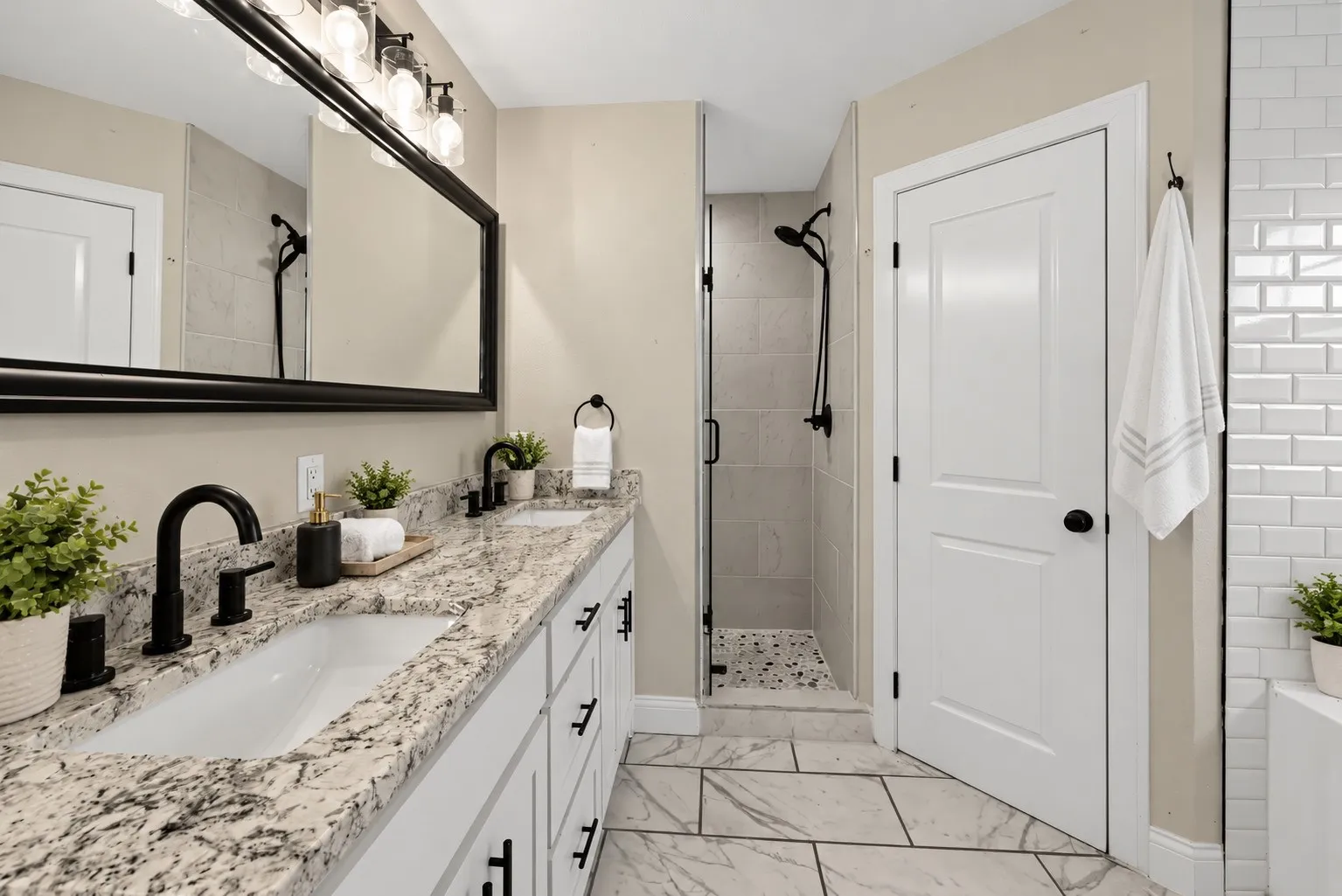 Dual-sink vanity with stone countertop and matte black fixtures, full-width mirror with integrated lighting, glass-enclosed shower with tiled walls, and large-format tile flooring