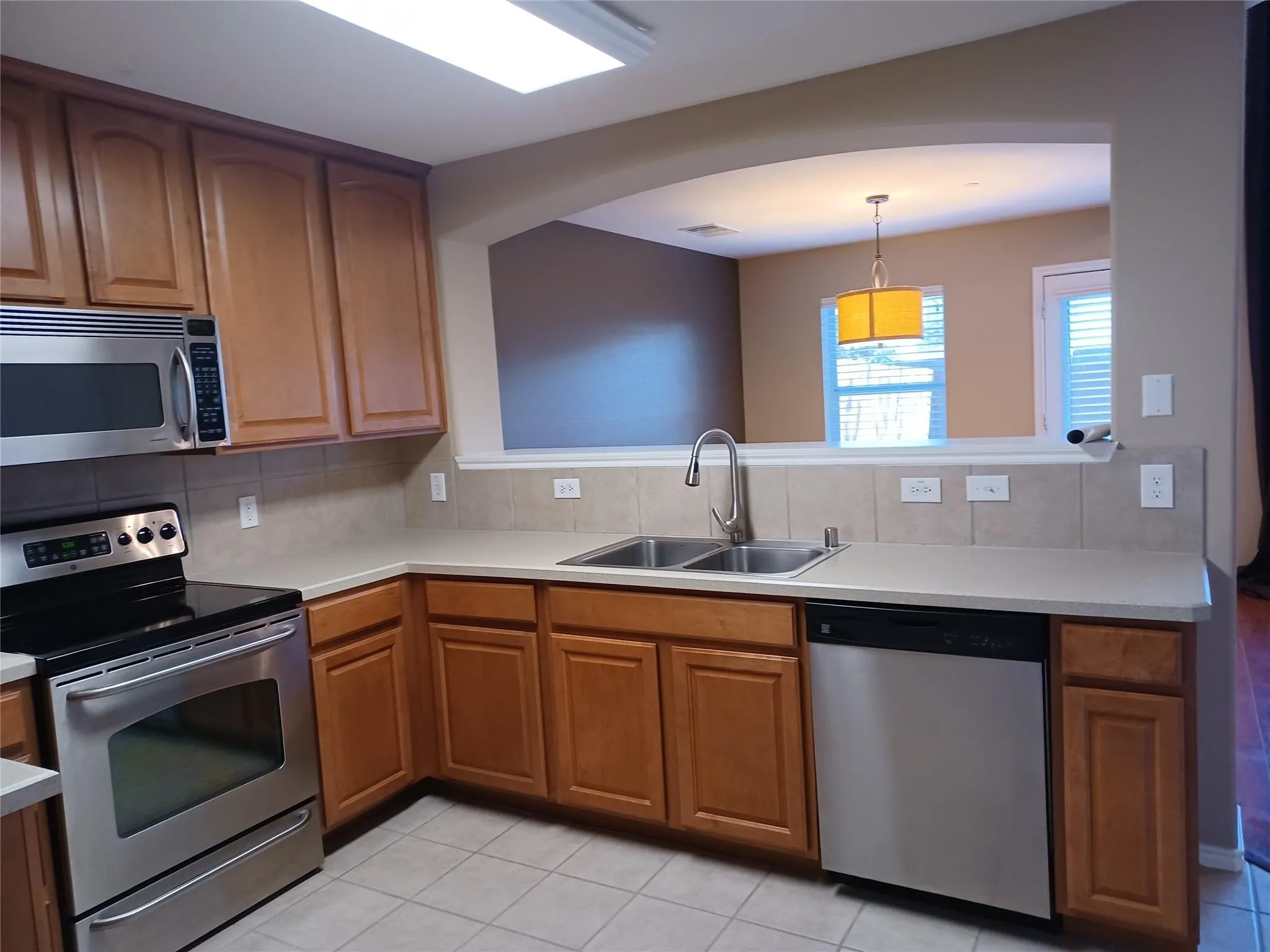 Kitchen featuring stainless steel appliances, wood-finish cabinetry, dual basin sink, light-toned countertops, and tile backsplash