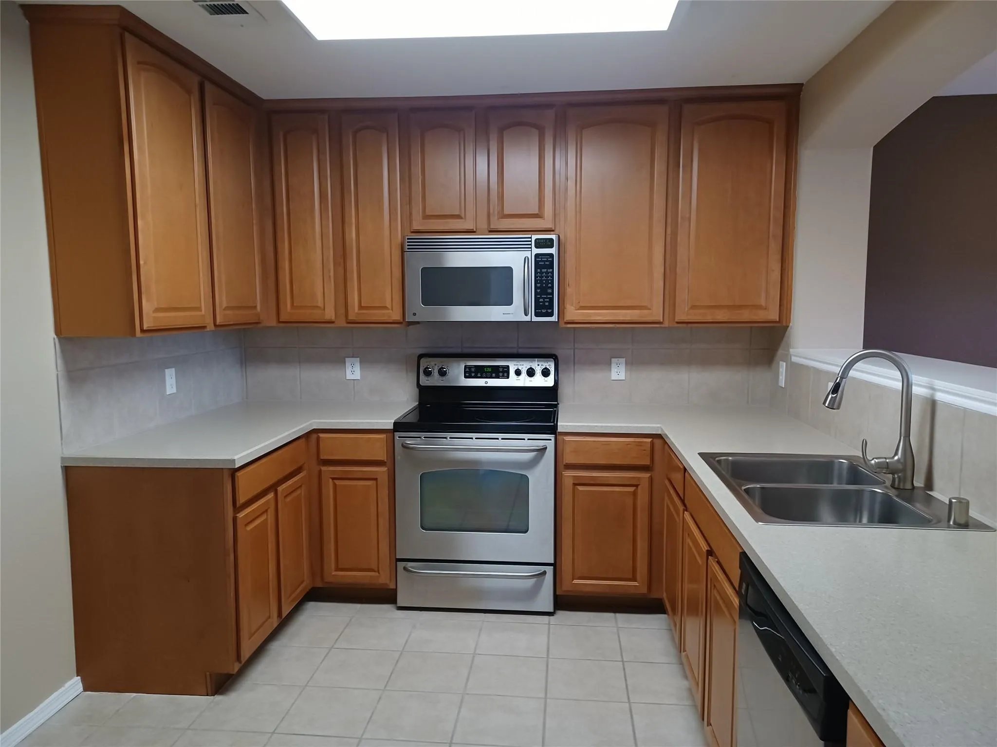 U-shaped kitchen featuring wood cabinetry, stainless steel appliances, light-toned countertops, tile backsplash, and tile flooring