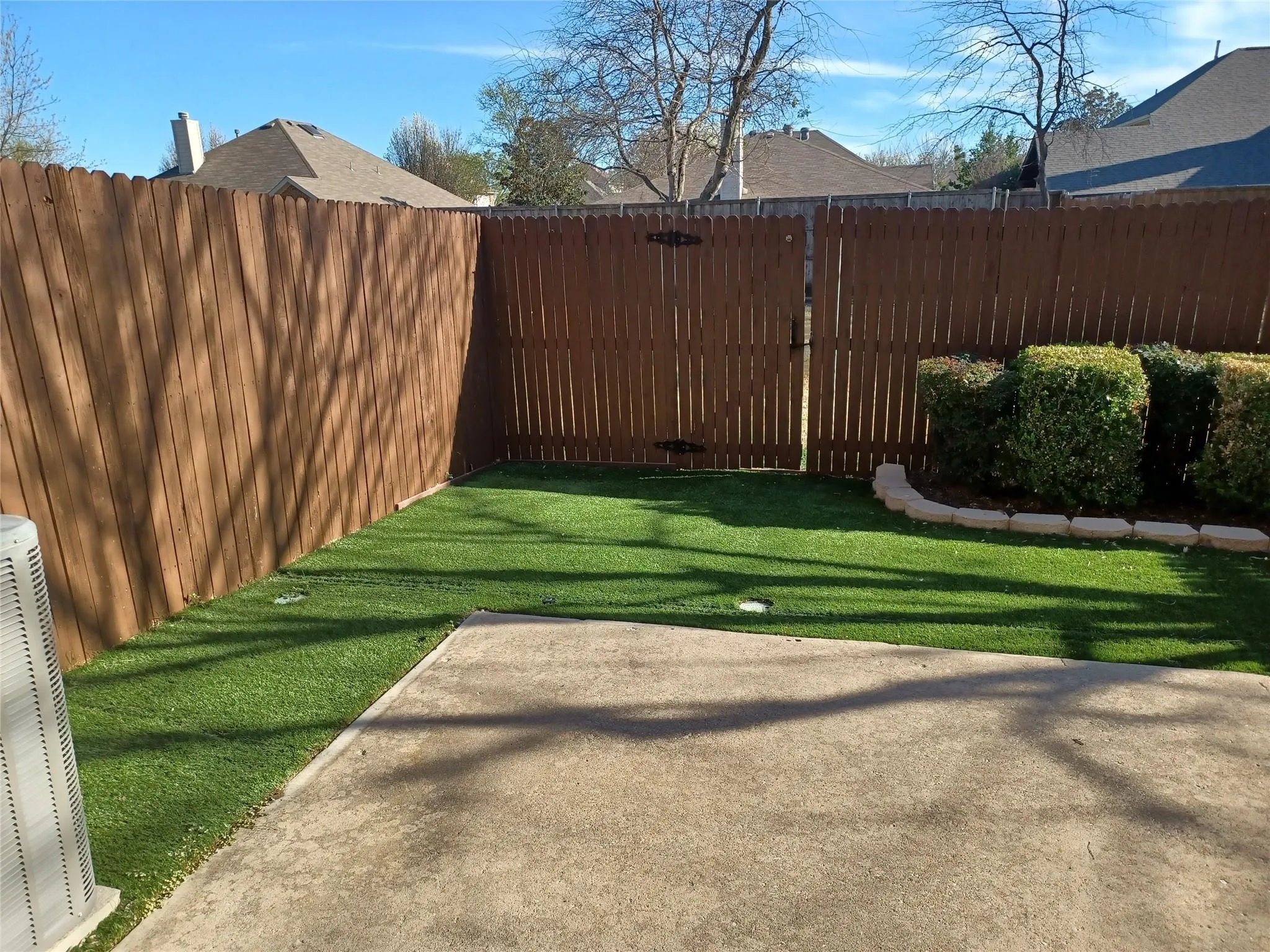 Fenced backyard featuring artificial turf, concrete patio, and landscaped shrubs with stone edging