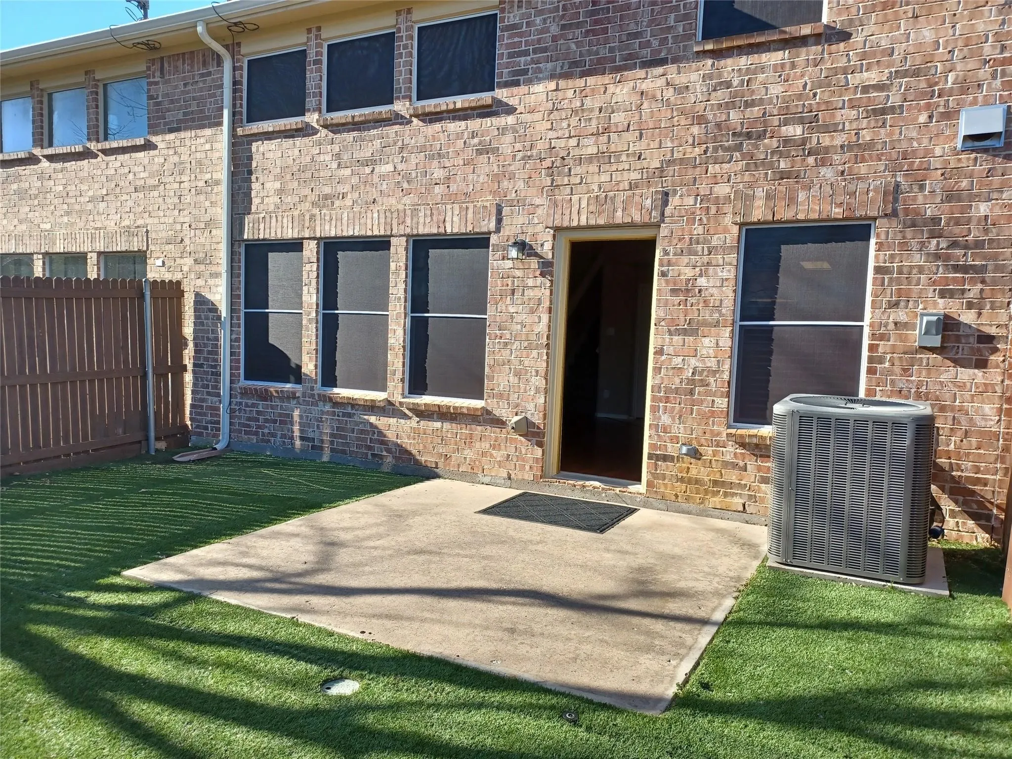 Brick exterior with multiple windows, a concrete patio, synthetic turf, and a wood privacy fence