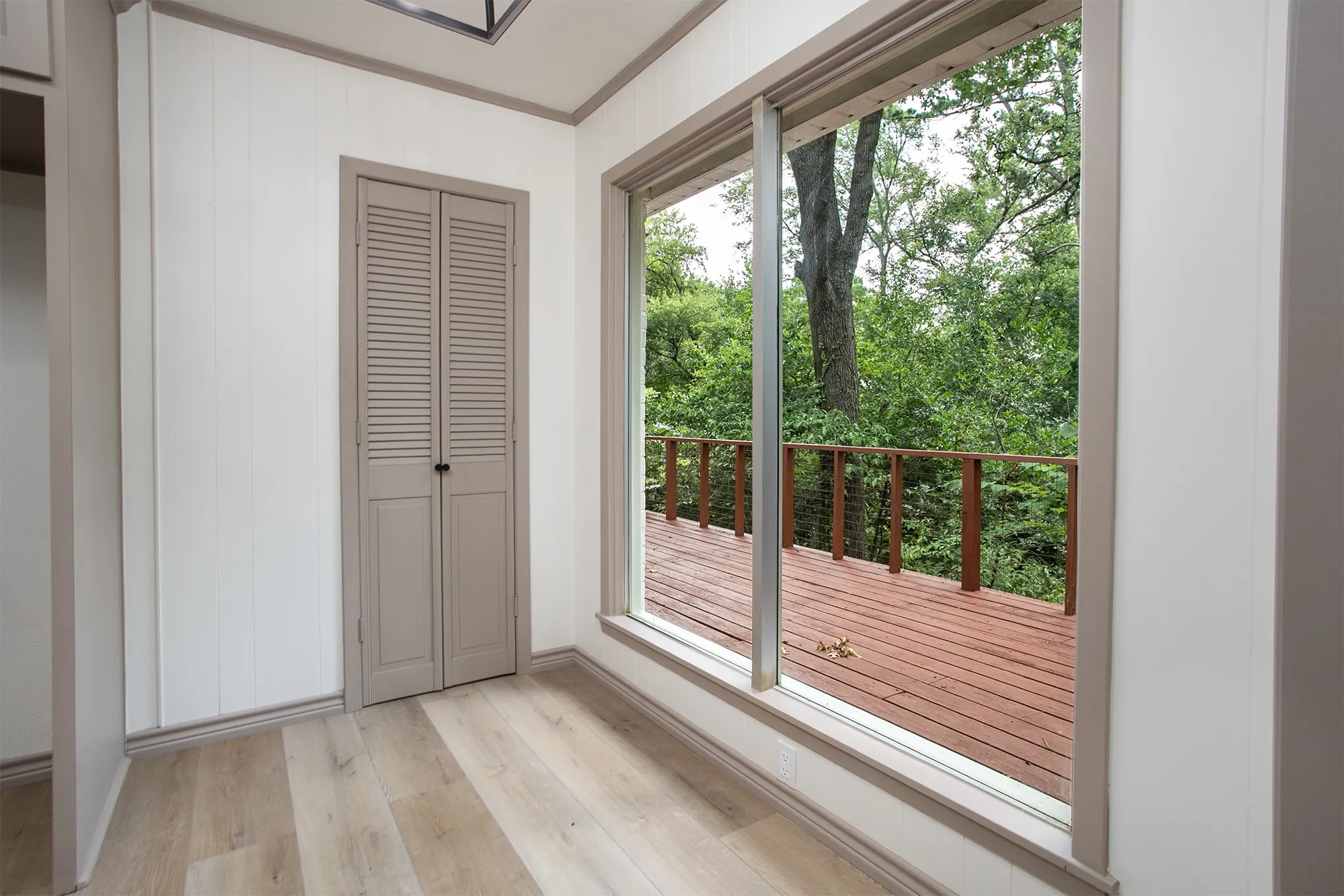 Wood-finish flooring, vertical paneling, louvered closet doors, and a large floor-to-ceiling window showcasing an exterior wood deck
