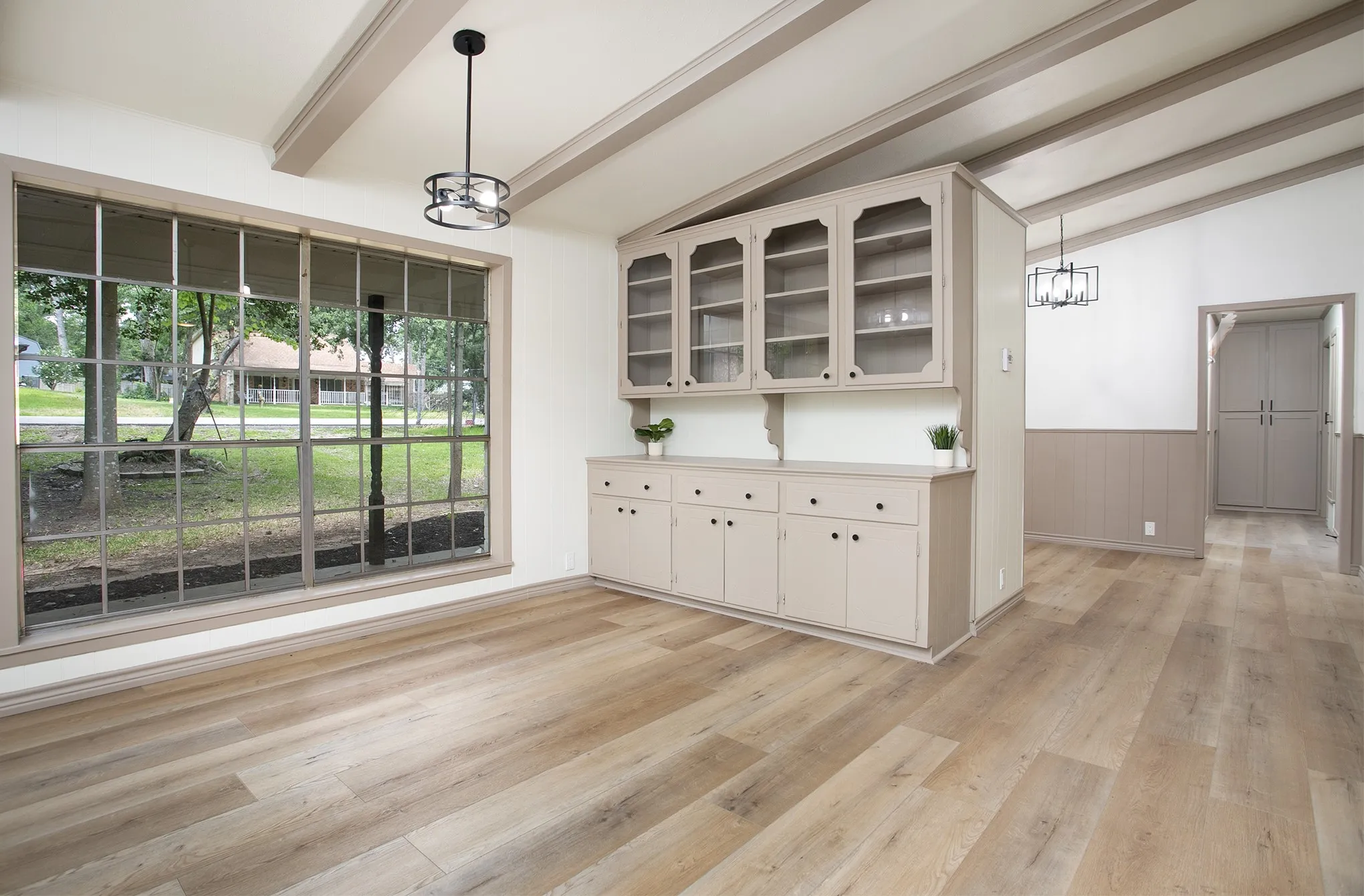 Large grid-pattern window, built-in cabinetry with glass-front display uppers, exposed ceiling beams, wood-finish flooring, and wainscoting