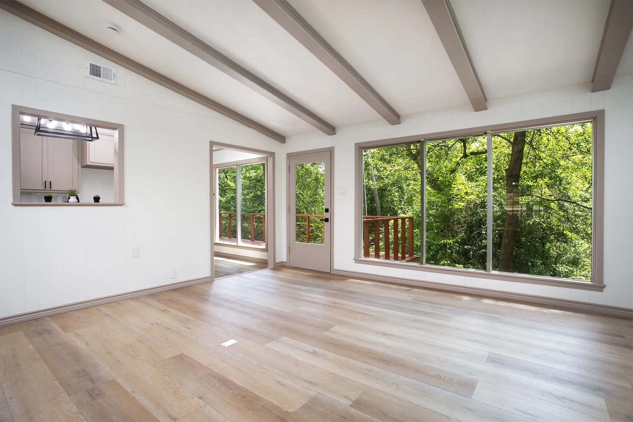 Living area featuring wood-finish flooring, exposed ceiling beams, expansive floor-to-ceiling windows, and a pass-through opening to the kitchen area