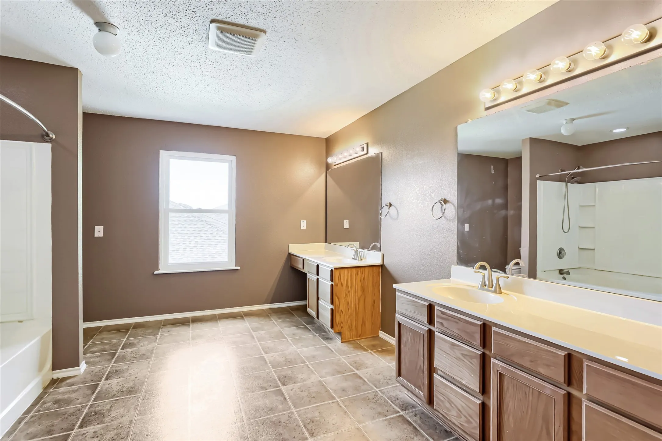 Dual-sink vanity with wood-finish cabinetry, ceramic tile flooring, full-width vanity mirrors, multi-bulb wall lighting, and tub-shower combination