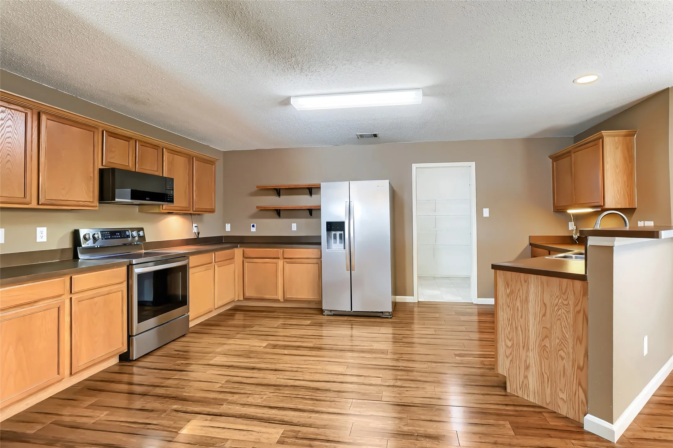 Kitchen featuring wood-finish cabinetry, stainless steel appliances, wood-finish flooring, floating wall shelving, and a peninsula counter